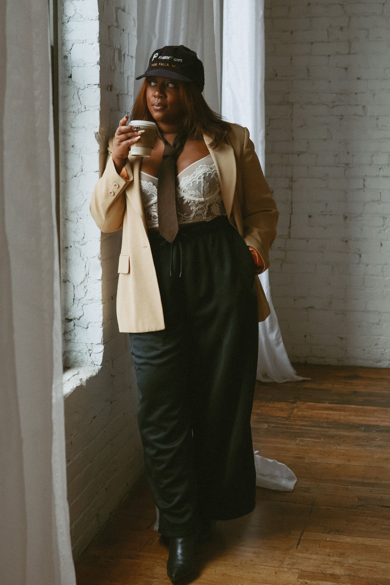 Stylish woman in tan blazer, lace top, and black pants holding coffee cup near bright window.