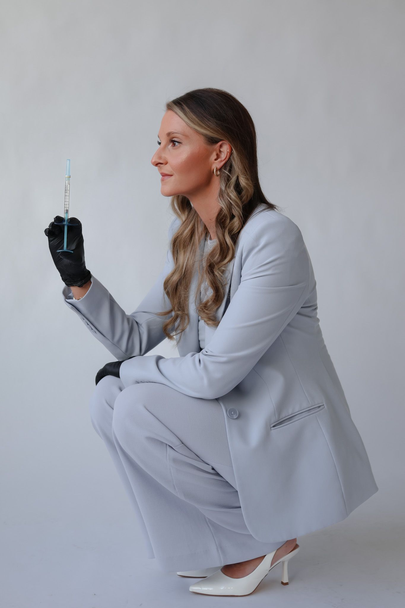 Blonde woman in grey suit crouching while holding medical syringe with black gloves on white background.