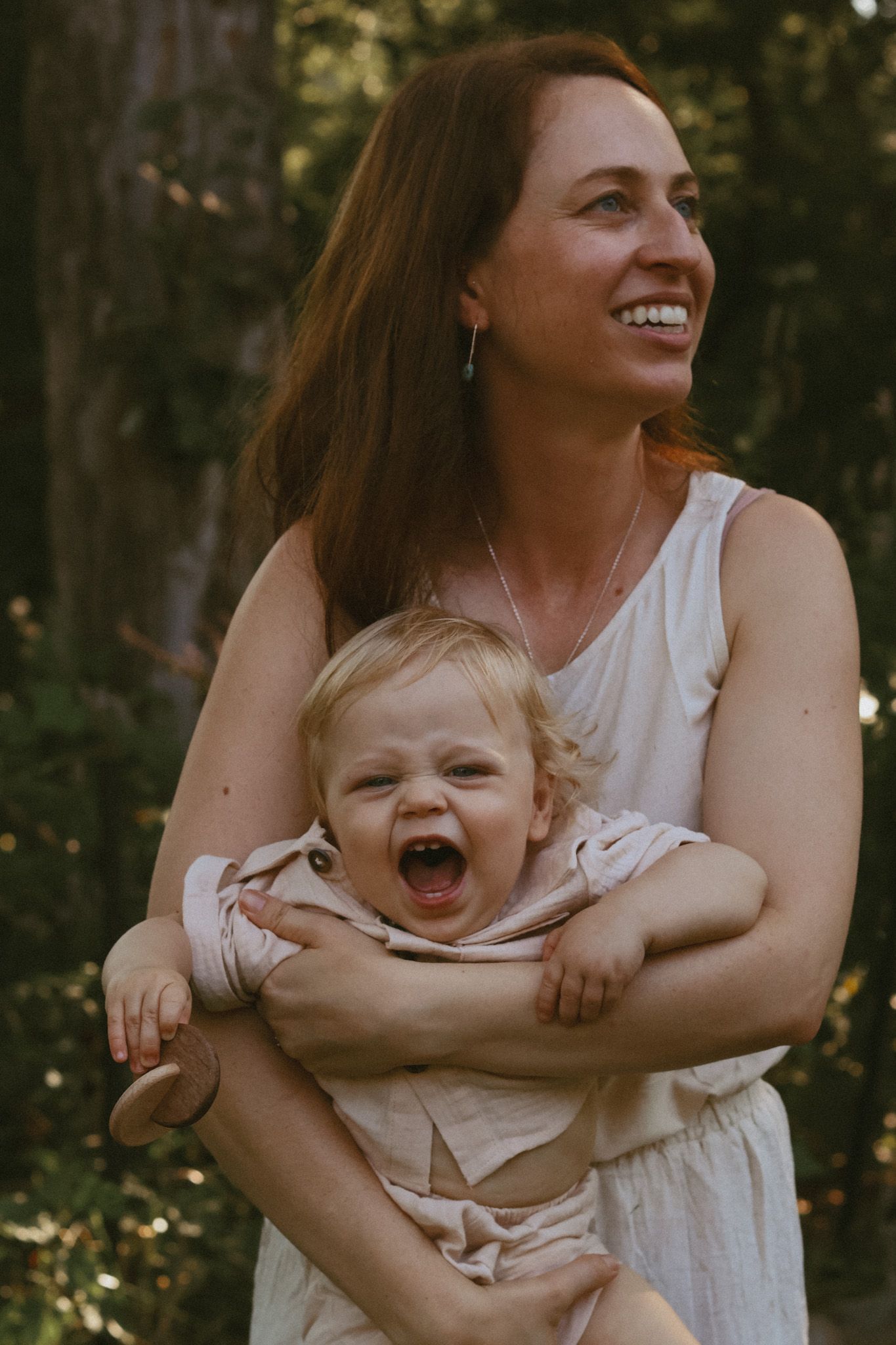 Smiling mother with long red hair holding a laughing yawning baby outdoors in a sunlit forest setting.