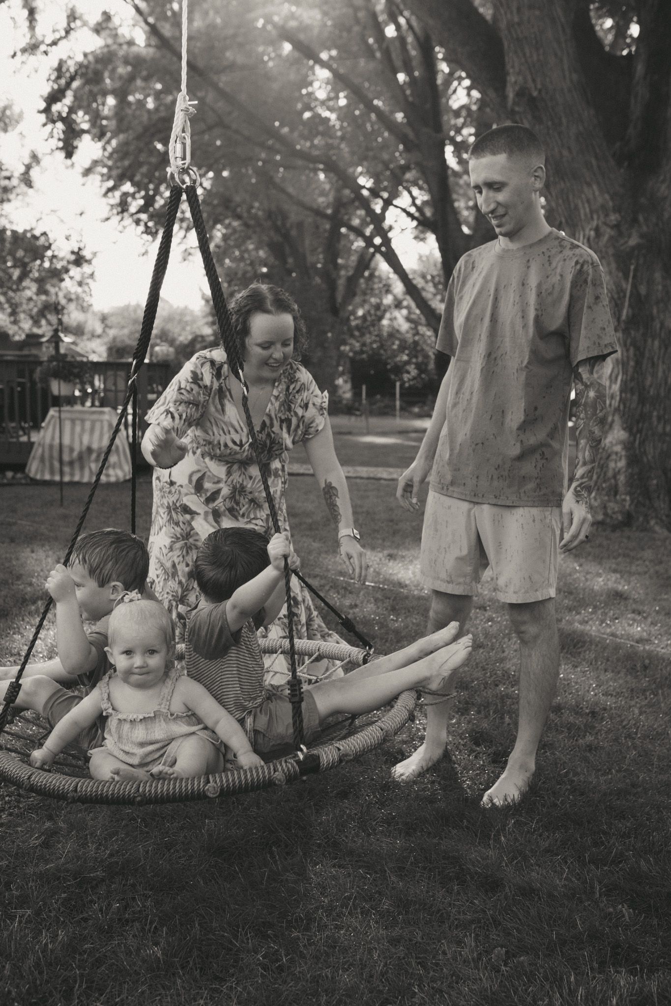 Black and white photo of a family with young children playing on a round rope swing in a backyard.