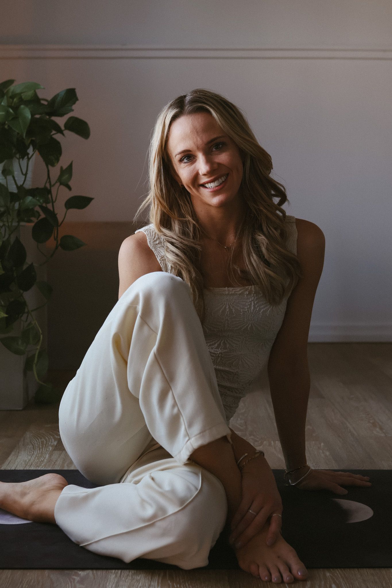 Smiling woman in white outfit sitting on yoga mat with green plant in background.