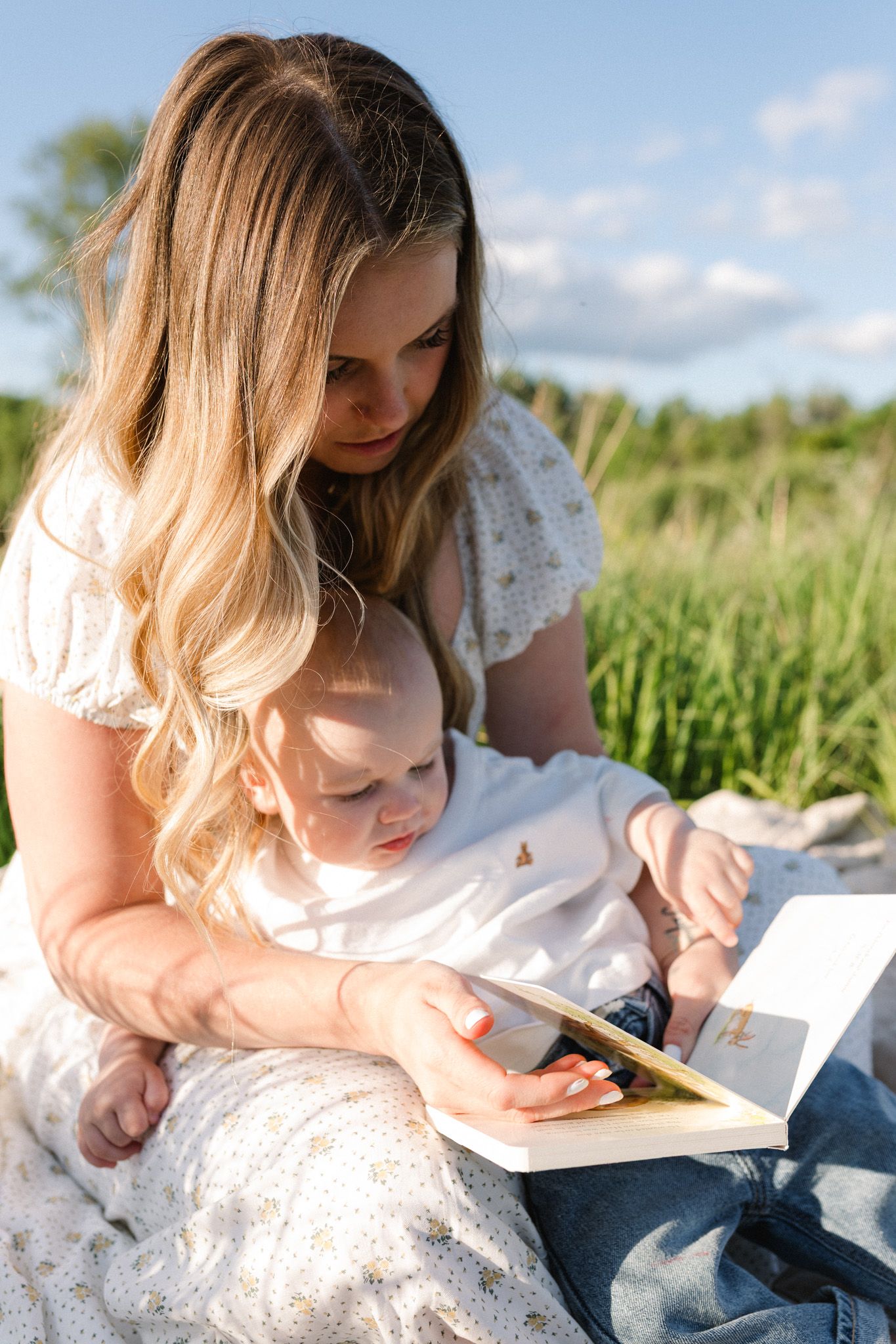 A mother in a white dress reads a book with her baby outdoors in a sunny green field.