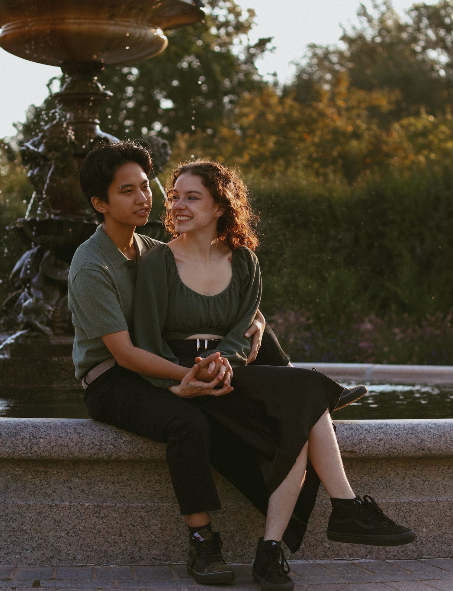 Happy young couple holding hands and sitting together by an ornate stone fountain in a sunny park.