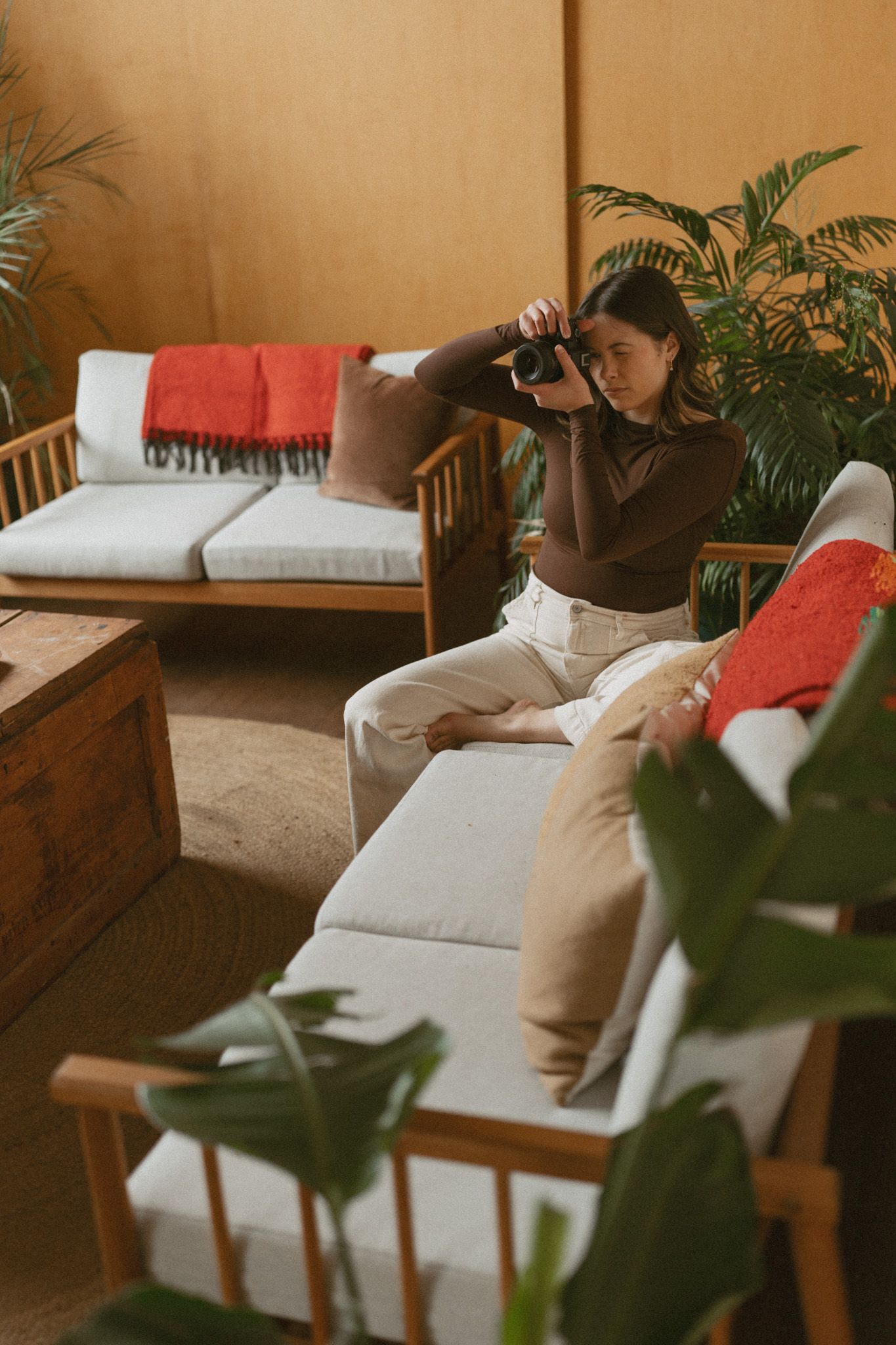 Young woman photographing with a film camera while seated in a warm boho living room with tropical plants.