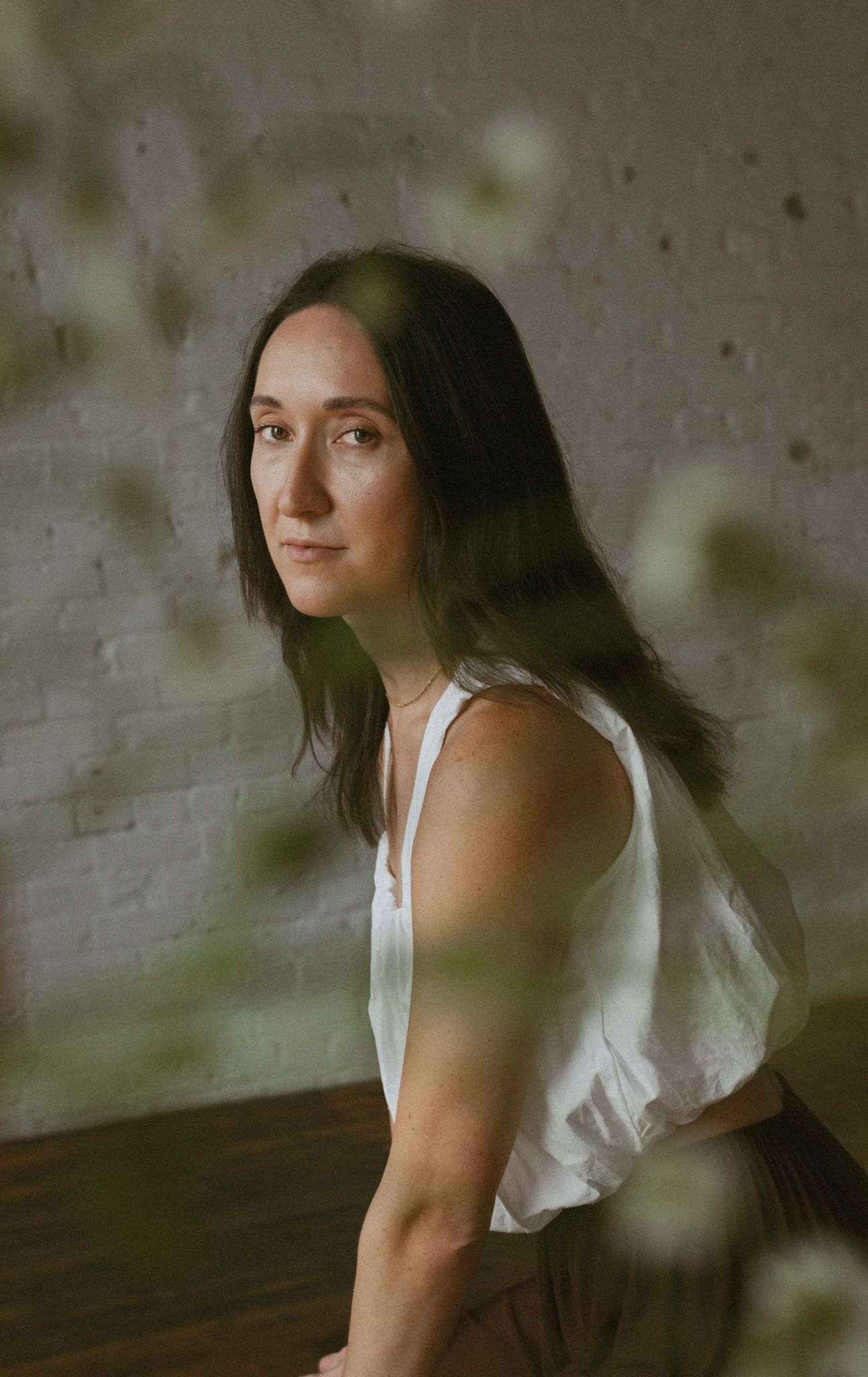 Dark-haired woman in white sleeveless top posing thoughtfully against a brick wall with soft foliage.