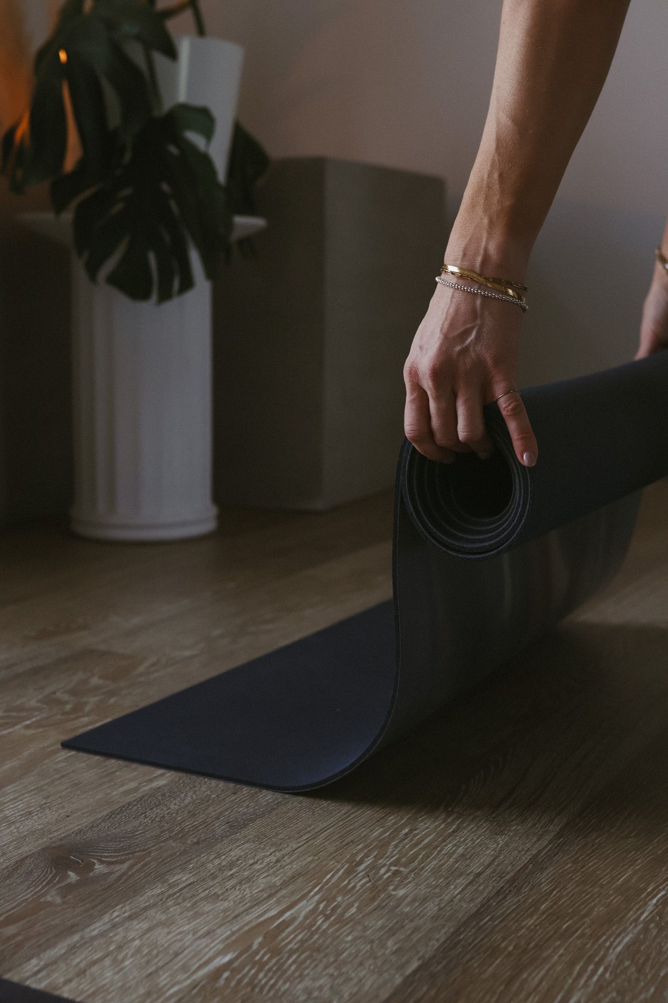 Hand with gold bracelet rolling up a dark yoga mat on hardwood floor.