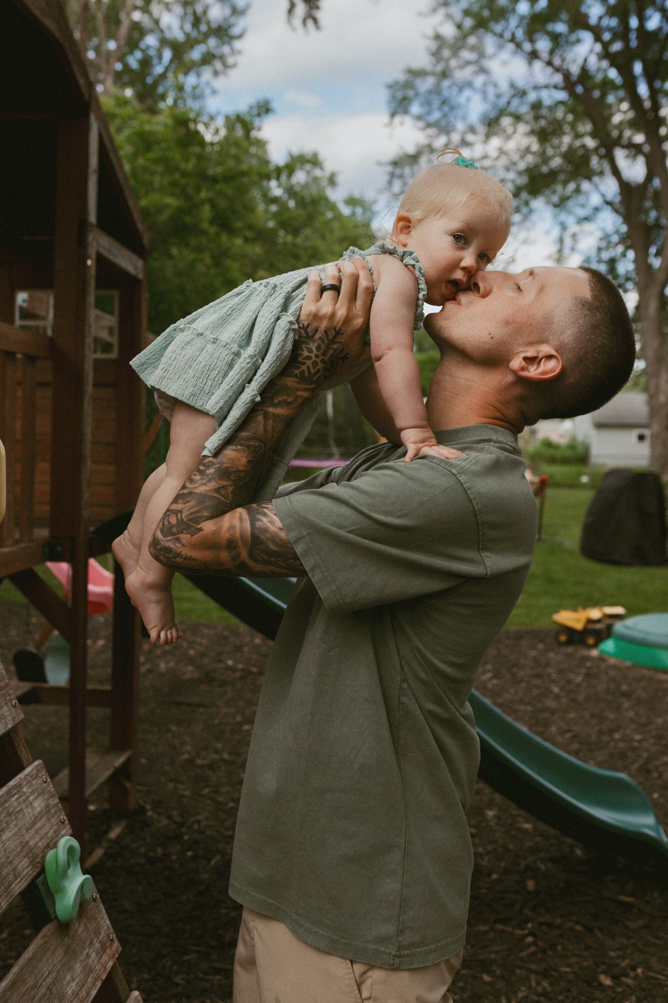 Tattooed father lifting and kissing his baby girl in a backyard playground setting.