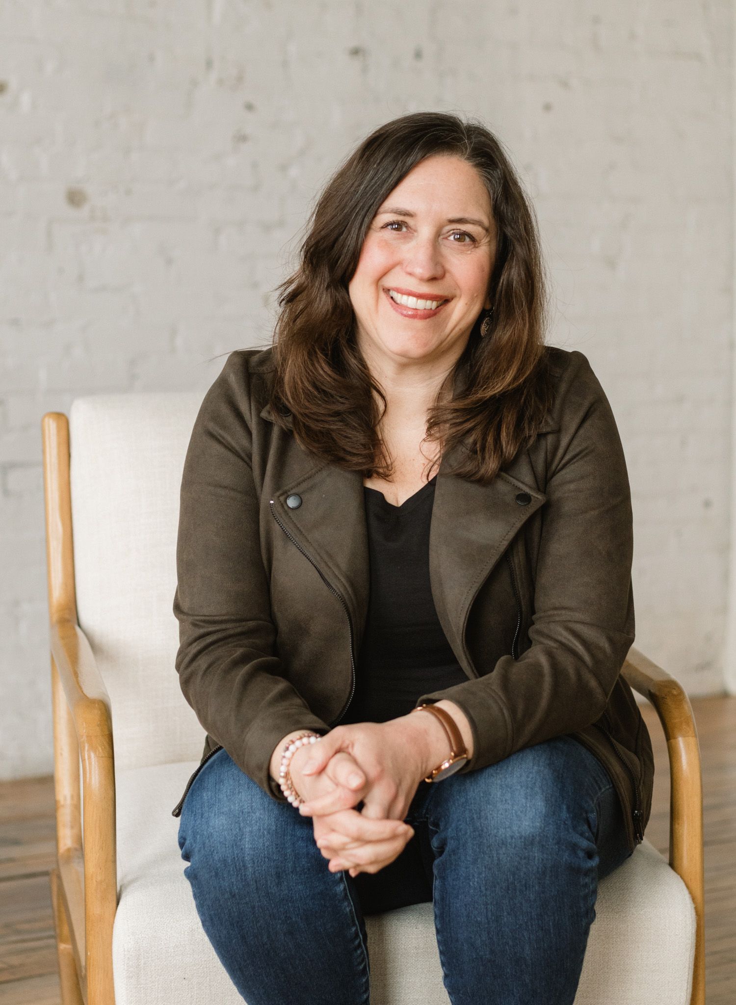 Smiling woman with brown hair wearing a olive jacket and jeans, sitting relaxed in a wooden chair indoors.