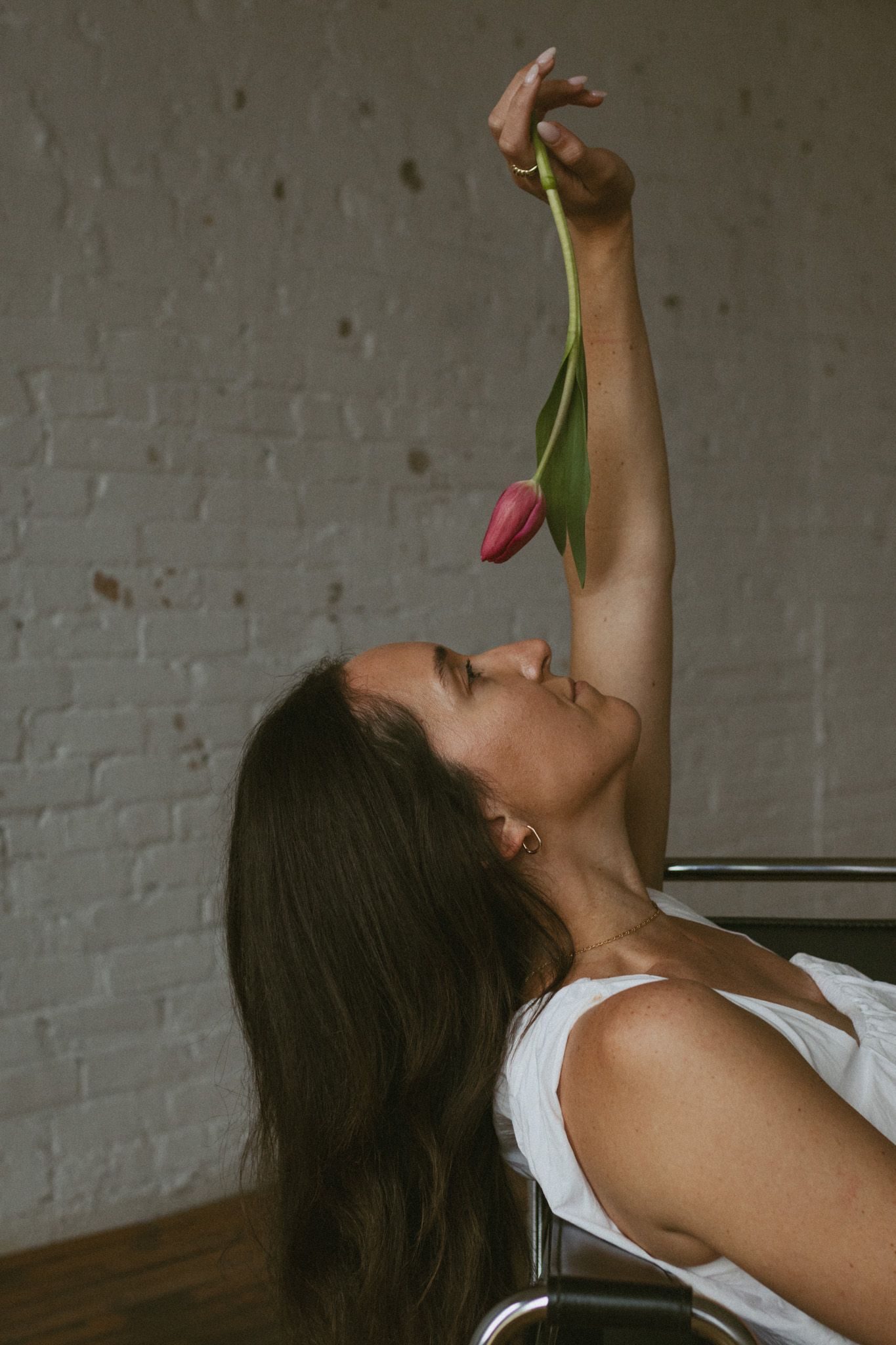 Woman reclining holding a pink tulip above her face against a white brick wall.