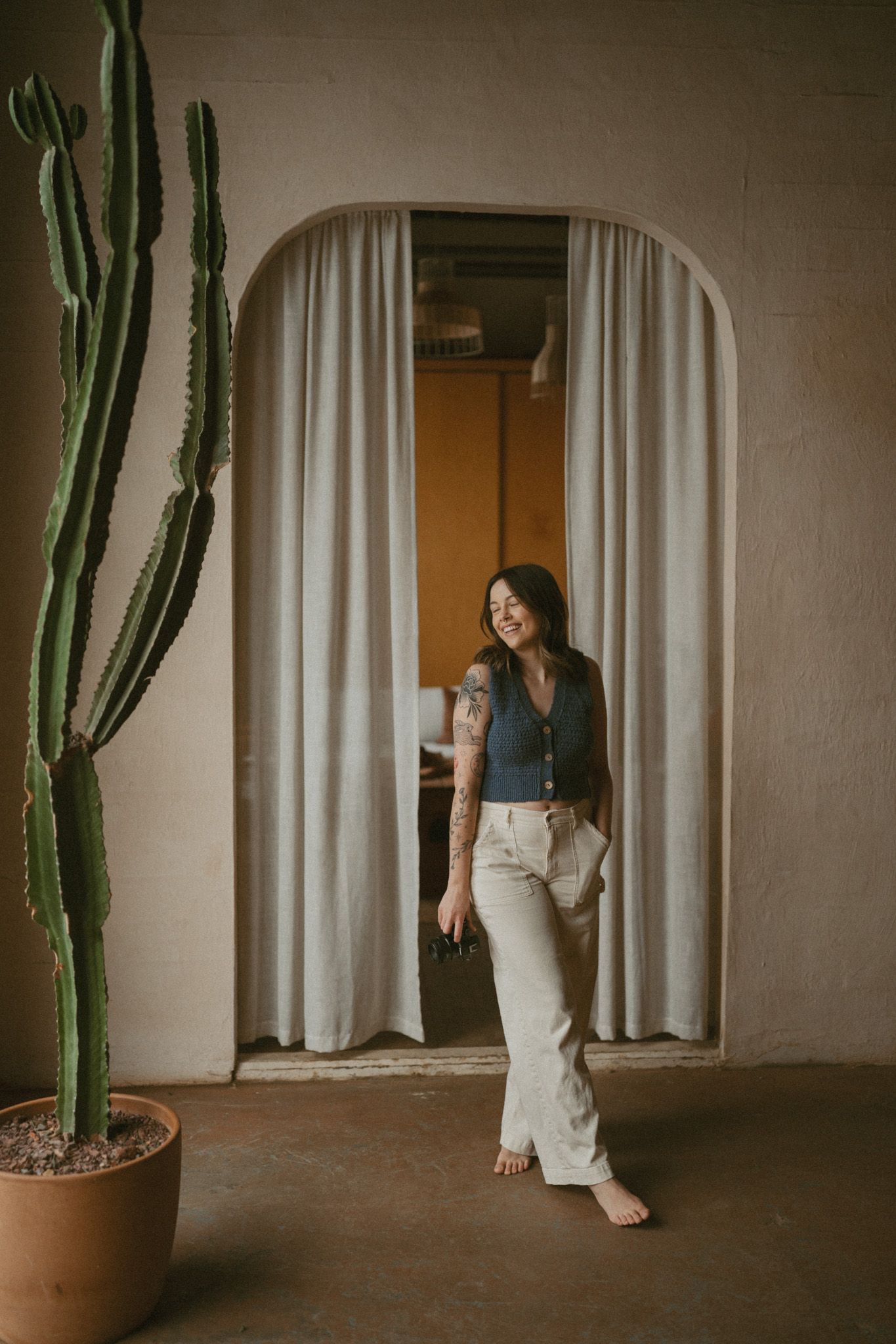 Smiling tattooed woman in wide-leg pants posing in arched doorway next to tall cactus.