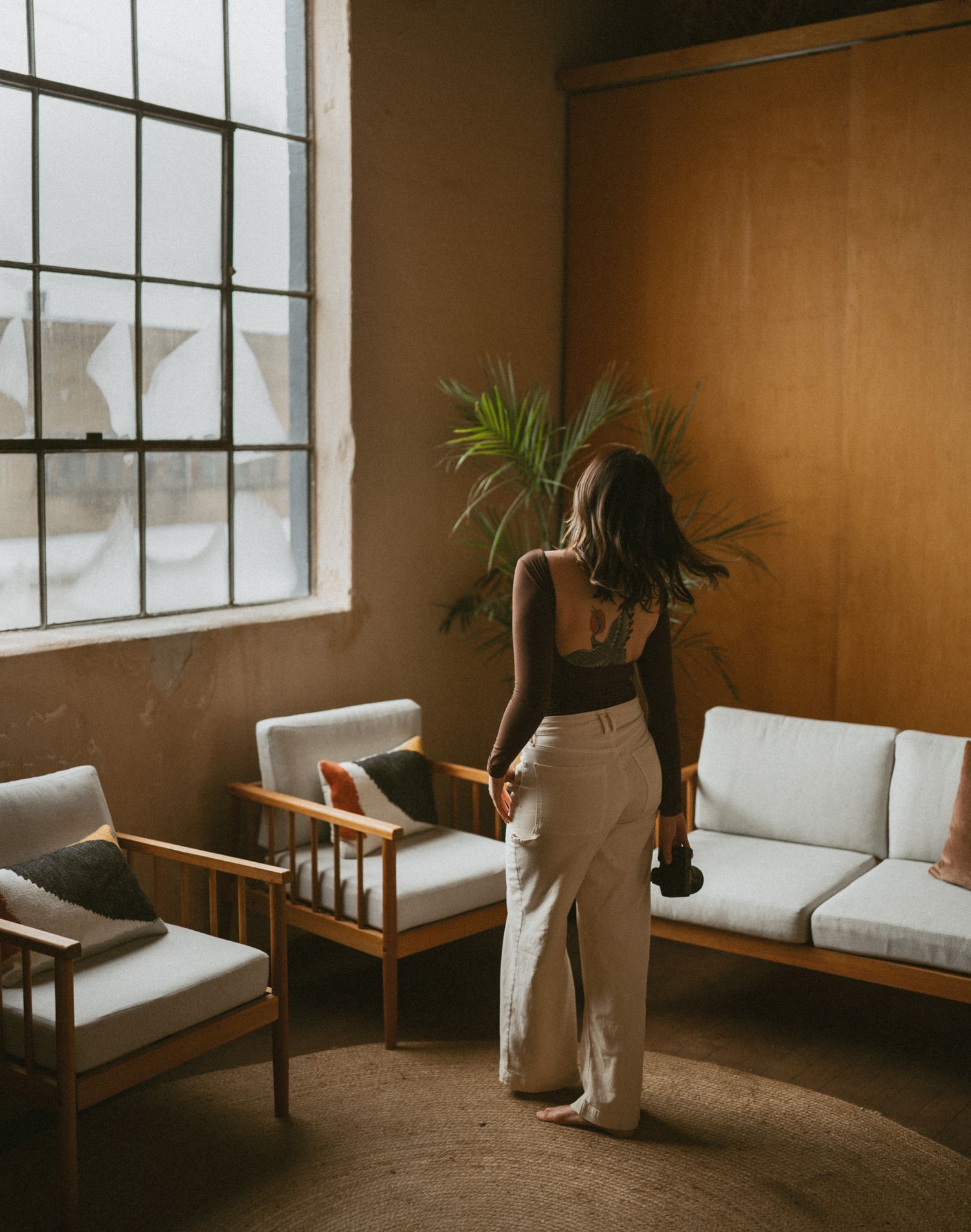 Woman in white maxi skirt with camera standing in warm sunlit lounge with wood paneling.