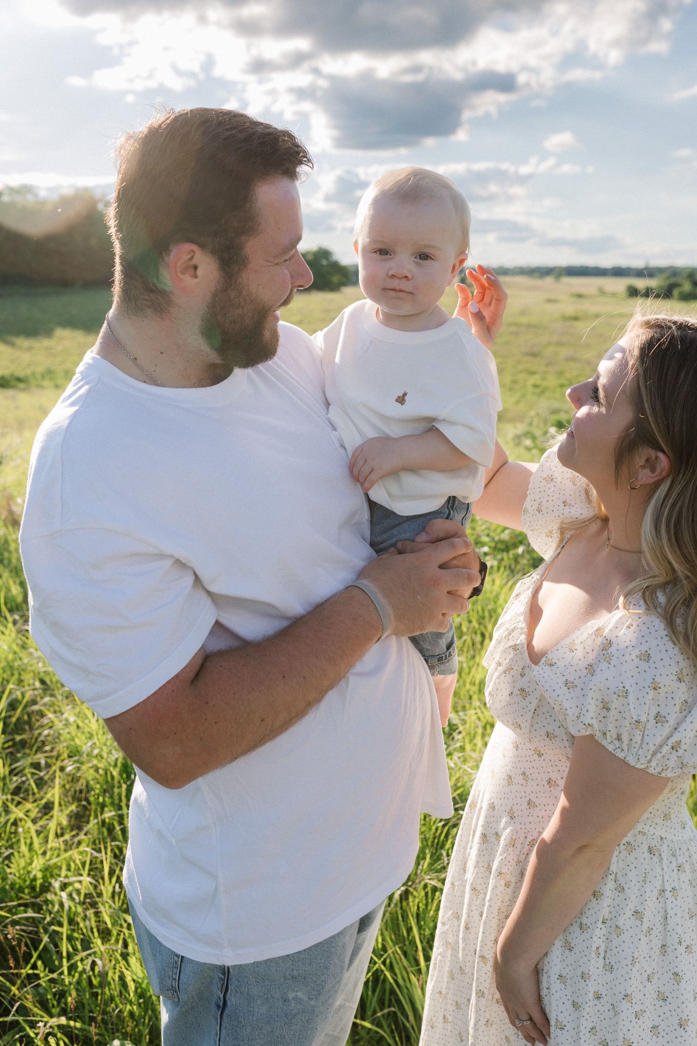 A bearded man in a white tee holds a baby while a woman in a floral dress smiles at them in a sunny green field.