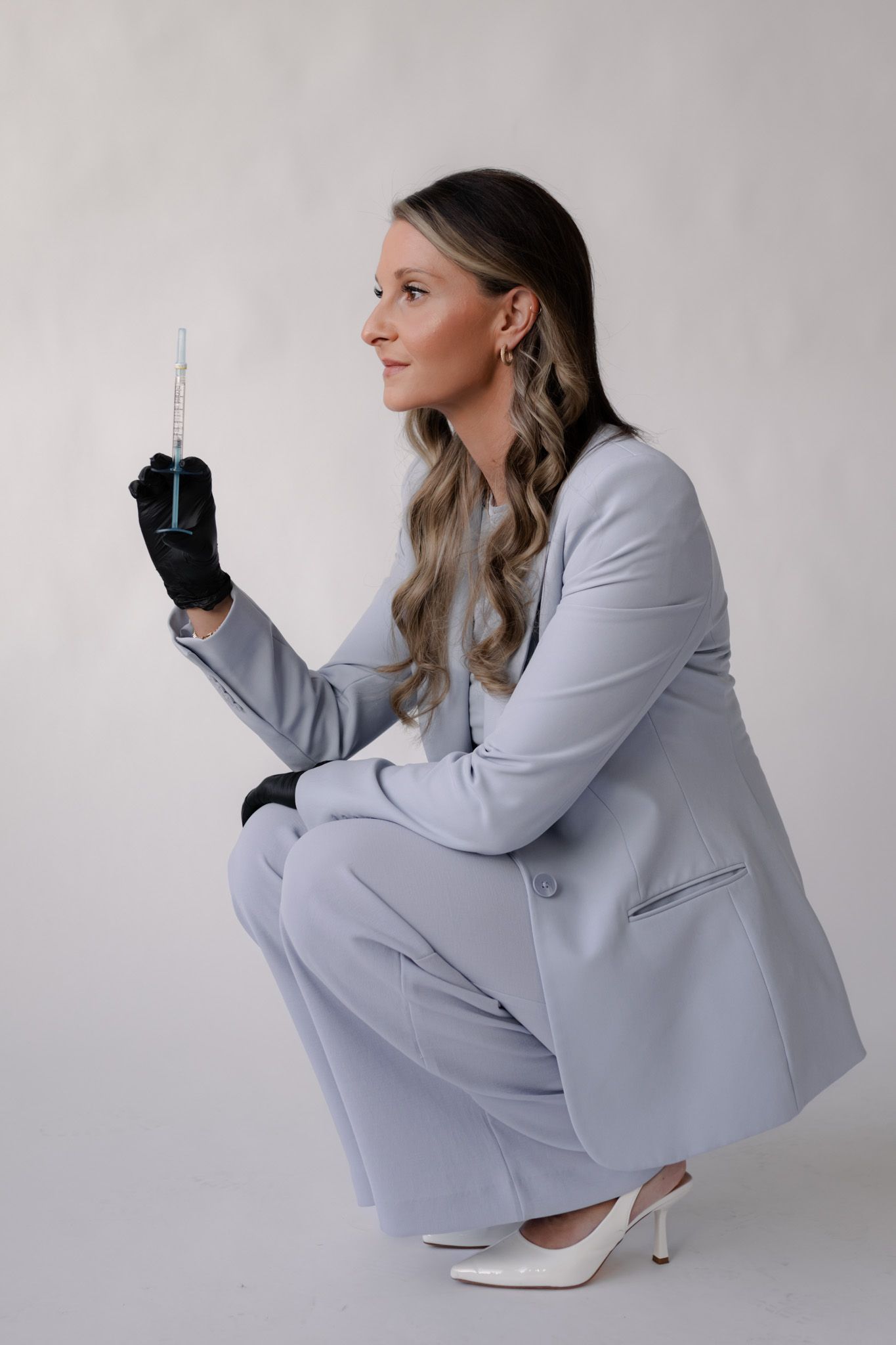 Woman in gray suit crouching while holding a syringe with black gloves.