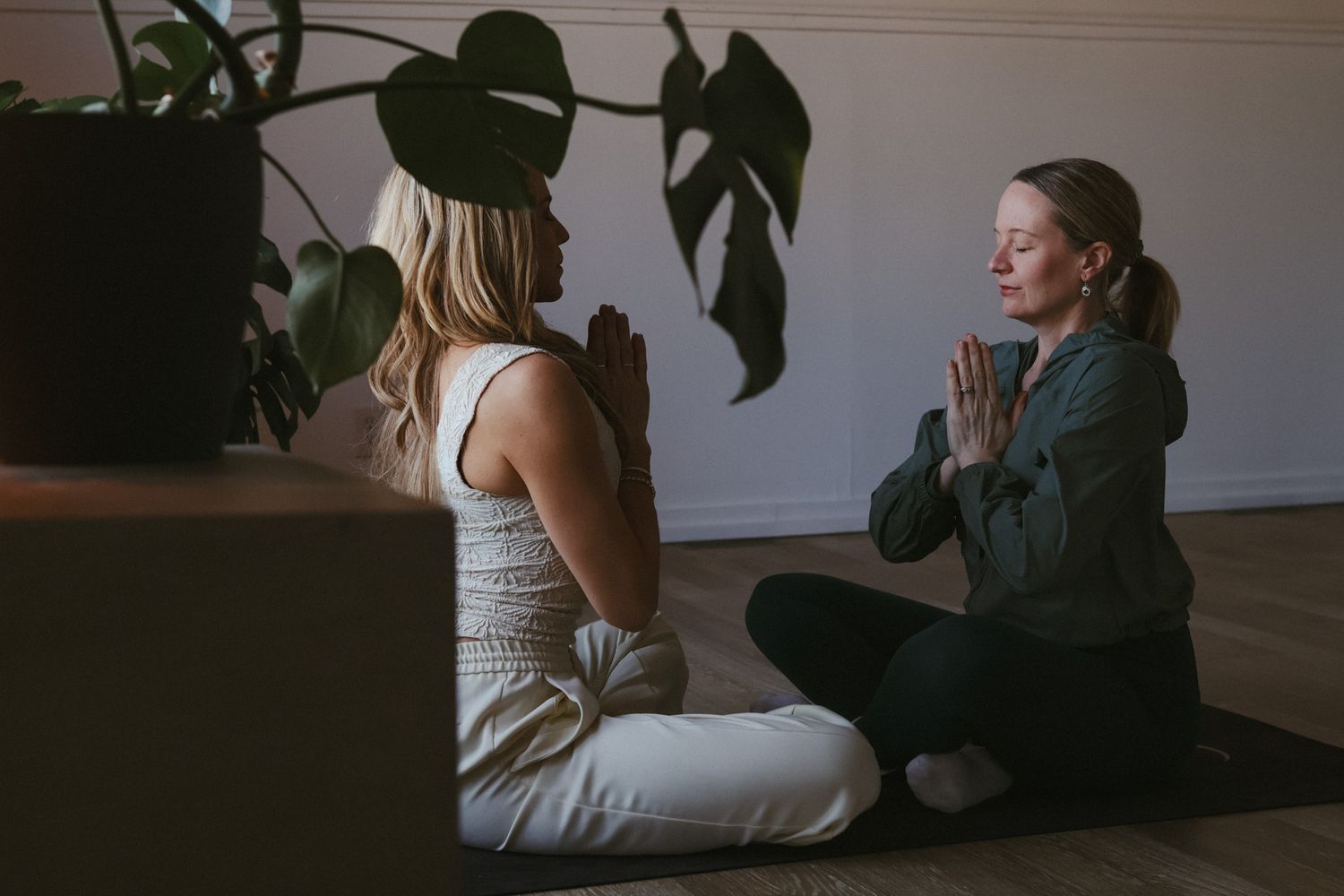 Two women sitting cross-legged on yoga mats facing each other in prayer pose during meditation session.