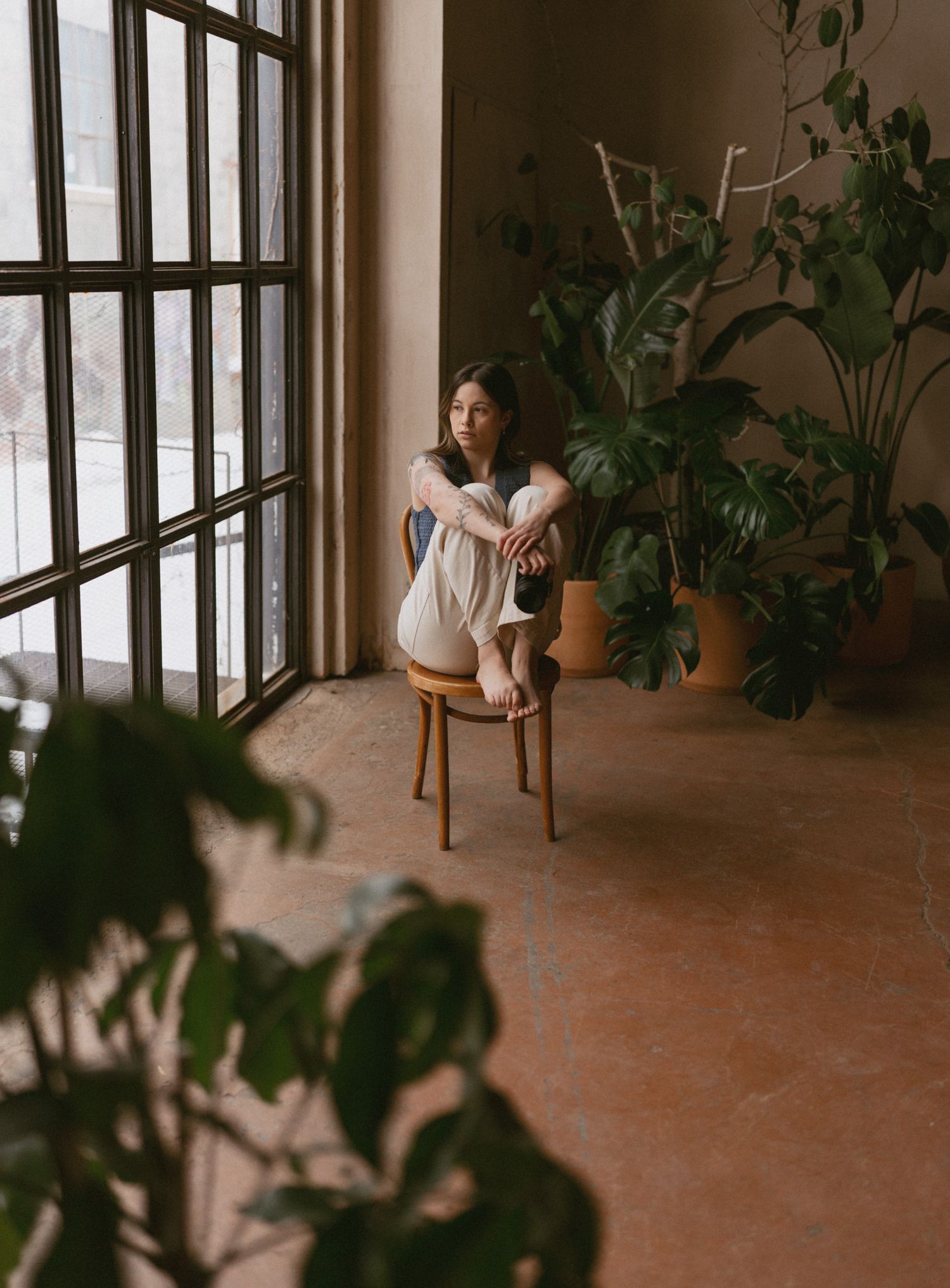 Young woman sitting curled up on a wooden chair beside large windows surrounded by lush green tropical plants.