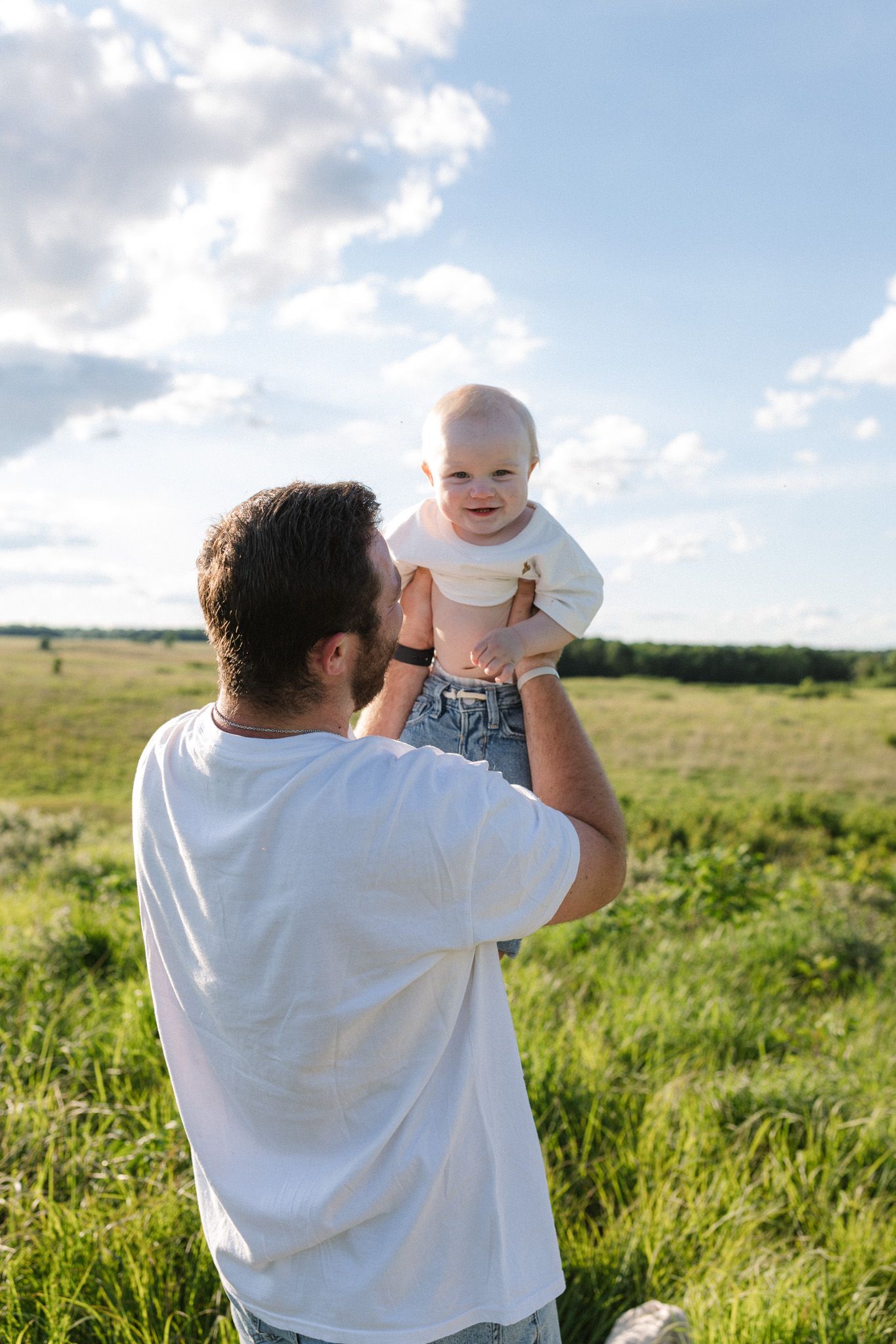 Father lifting happy baby in green field under blue sky, sharing a joyful outdoor moment together.