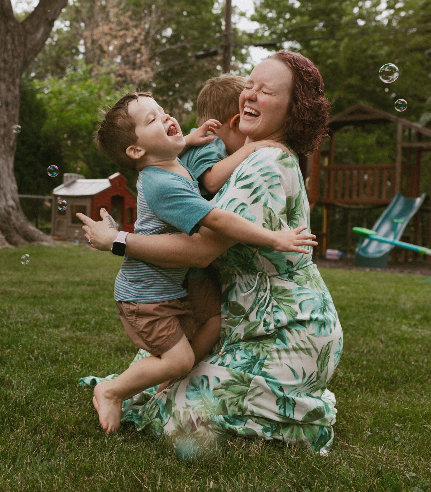 A joyful mother in a floral dress hugs two laughing boys outdoors on green grass with bubbles floating nearby.