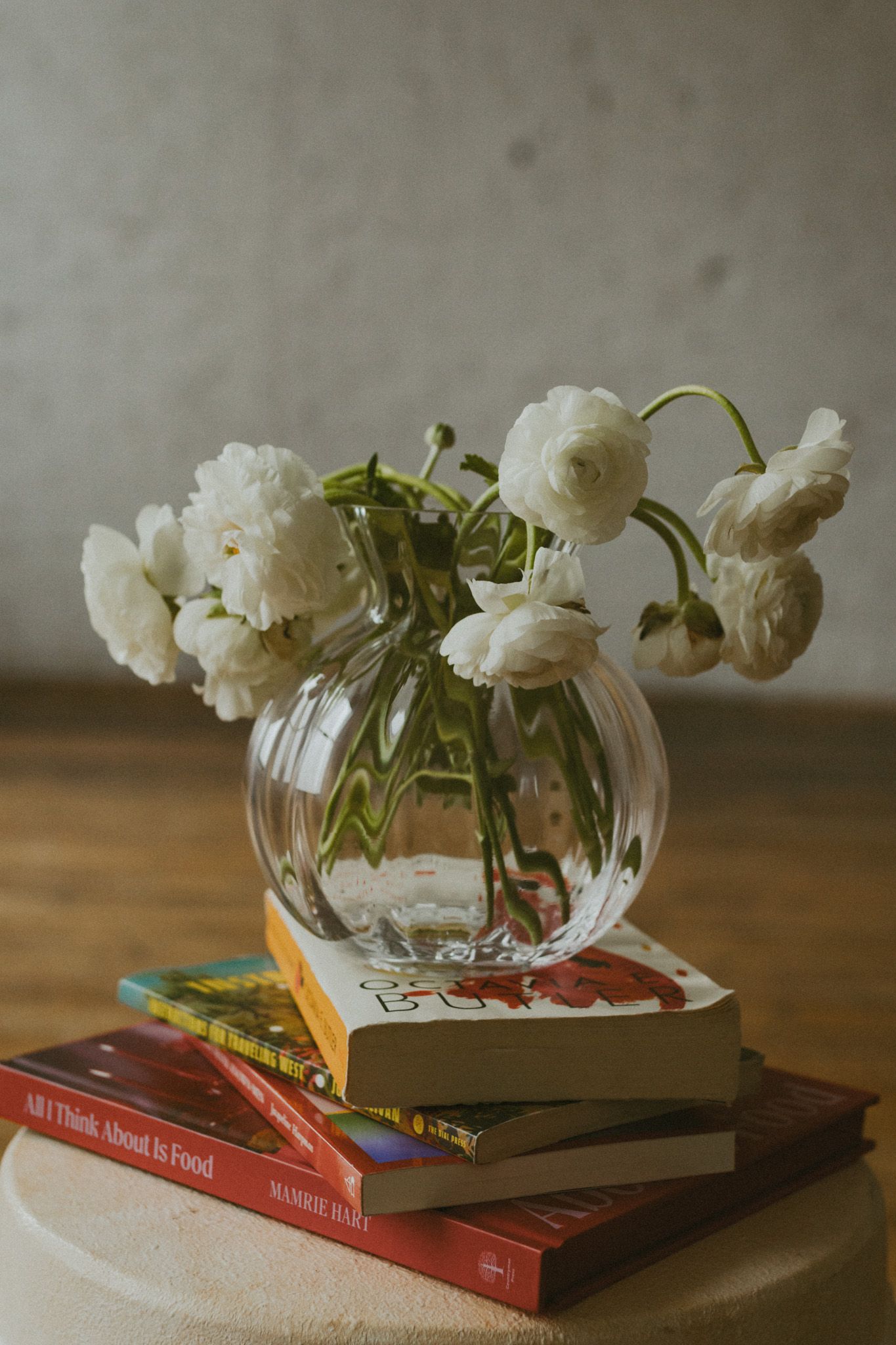 White ranunculus flowers in round glass vase atop stacked books on wooden table.