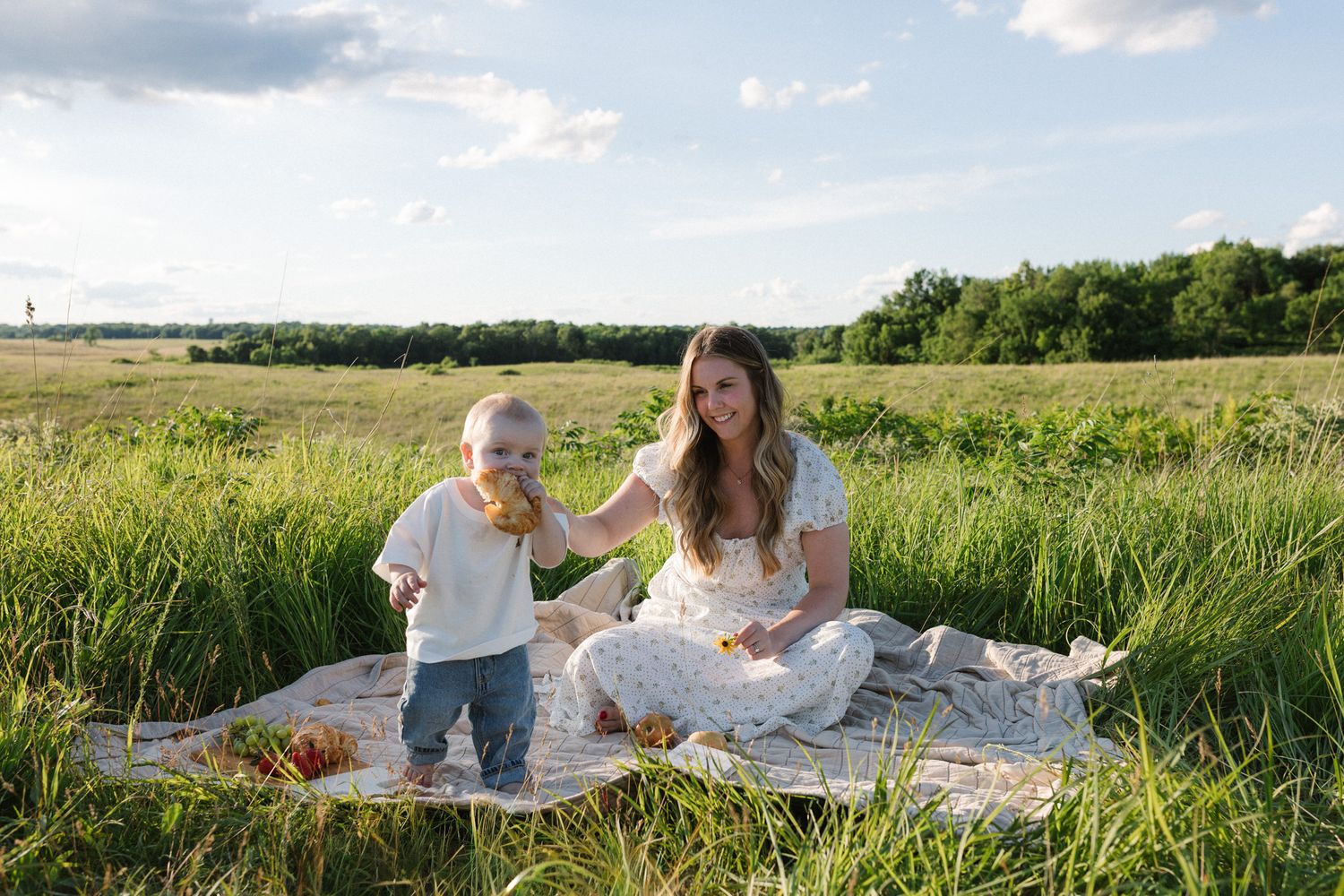 Mother and toddler enjoying a sunny outdoor picnic on a blanket in a lush green field on a beautiful day.