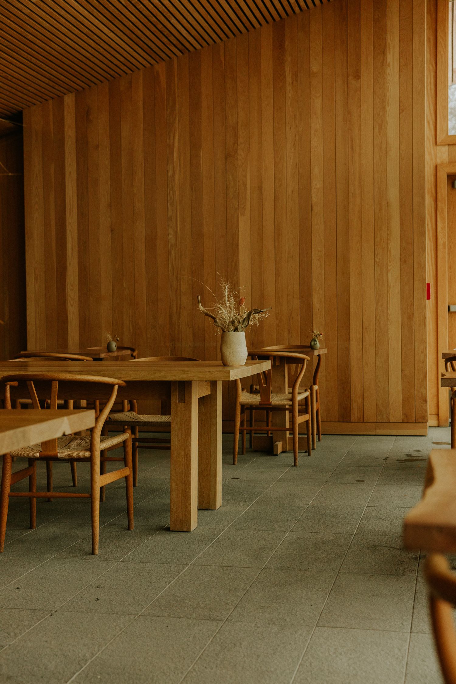 Minimalist dining room with wooden walls, wishbone chairs, and a light wood table with a dried flower vase.