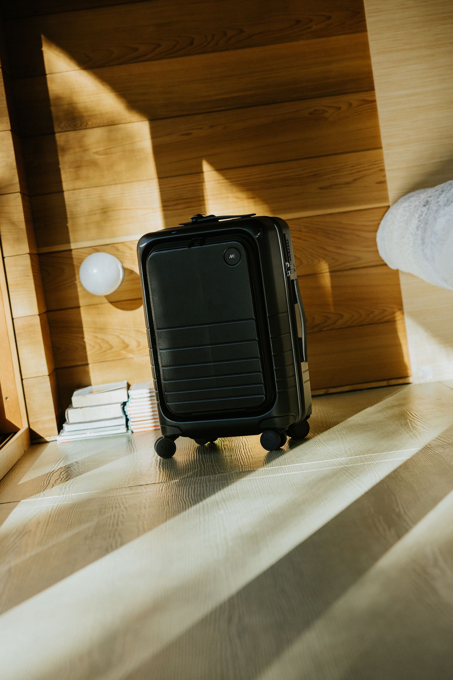 Black carry-on suitcase standing in sunlit wooden hotel room beside folded towels.