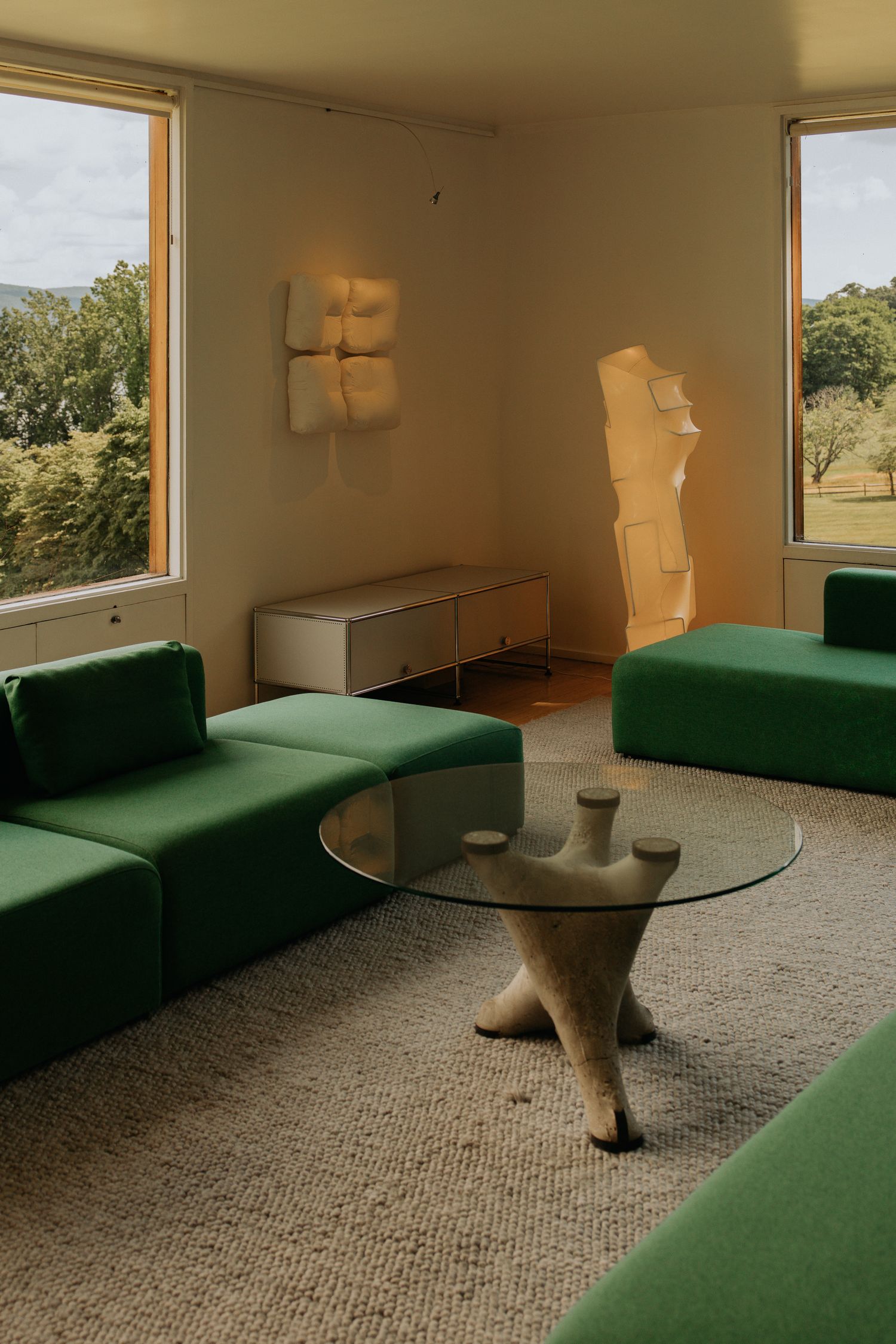 Mid-century living room with green modular sofas, glass table, and landscape windows.