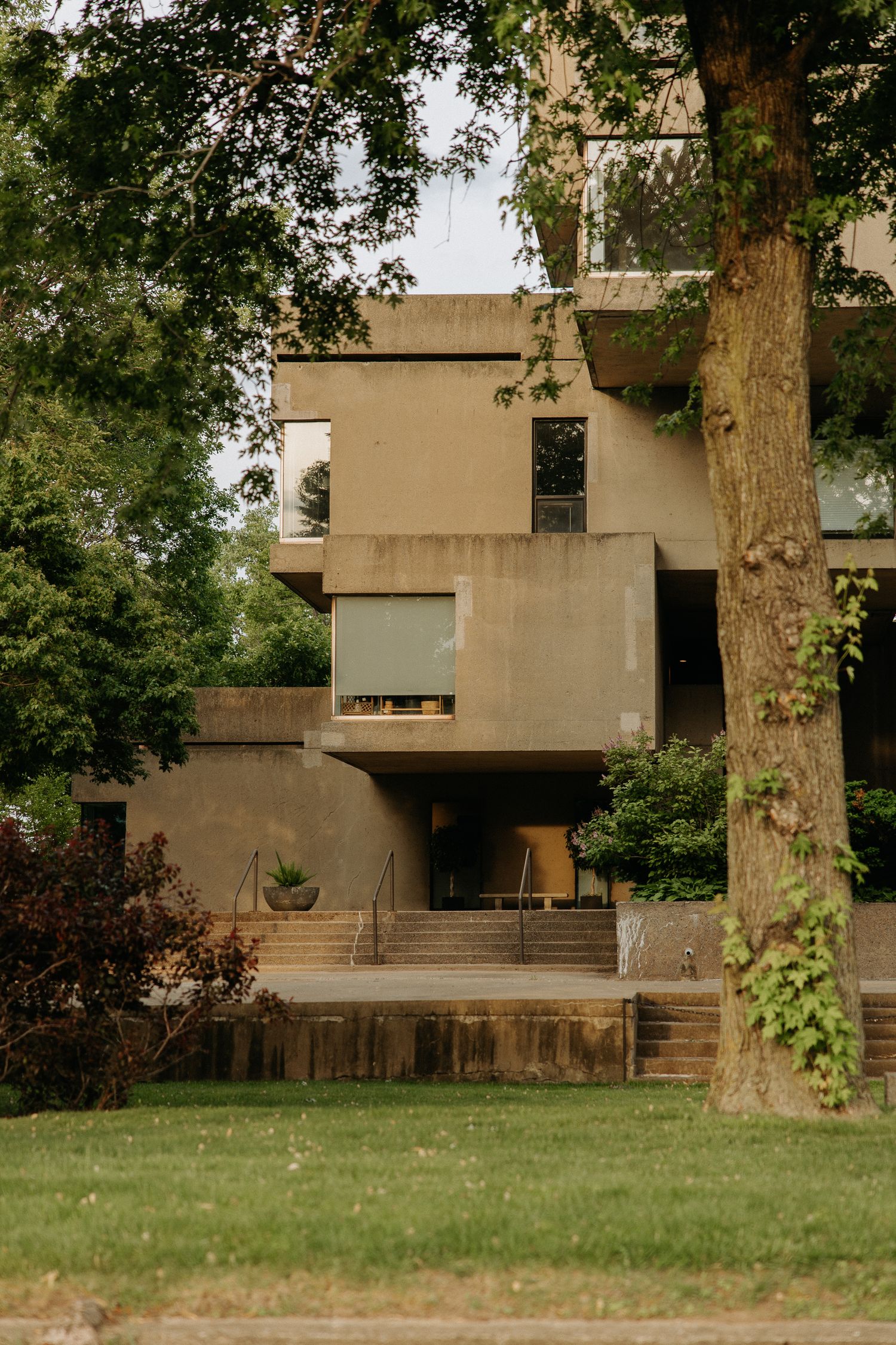 Brutalist concrete house surrounded by lush green trees and manicured lawn in a residential neighborhood.