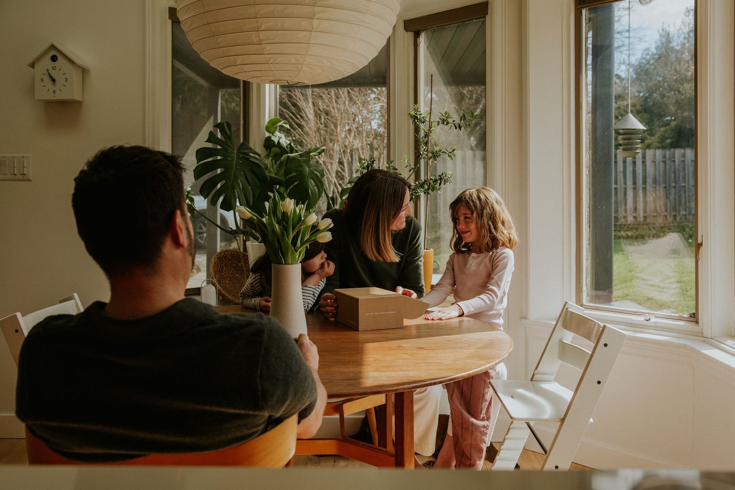 Family gathered around a round wooden dining table near bright sunlit windows with indoor plants.