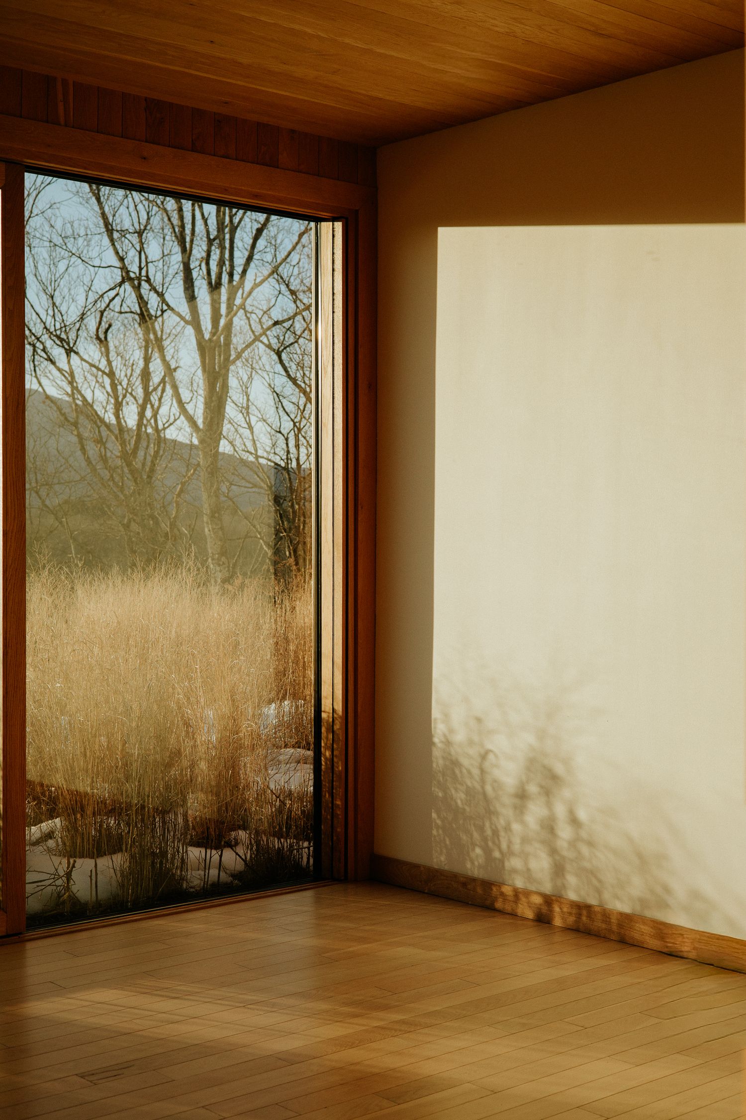 Minimalist interior room with wooden floors, large glass door overlooking dry autumn grasses, bare trees, and misty mountains.