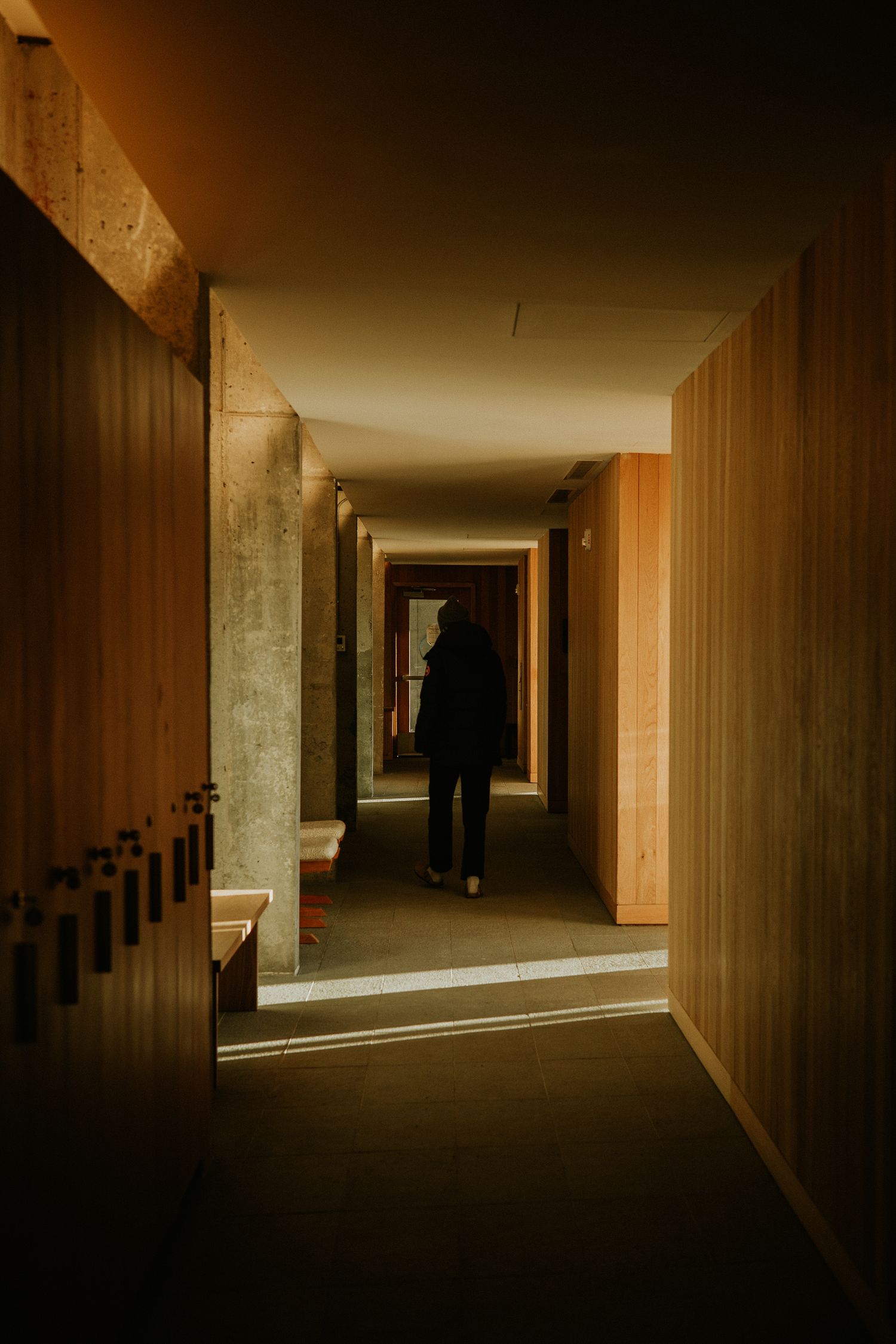A lone silhouette walks down a dimly lit corridor with warm wooden walls and concrete accents.