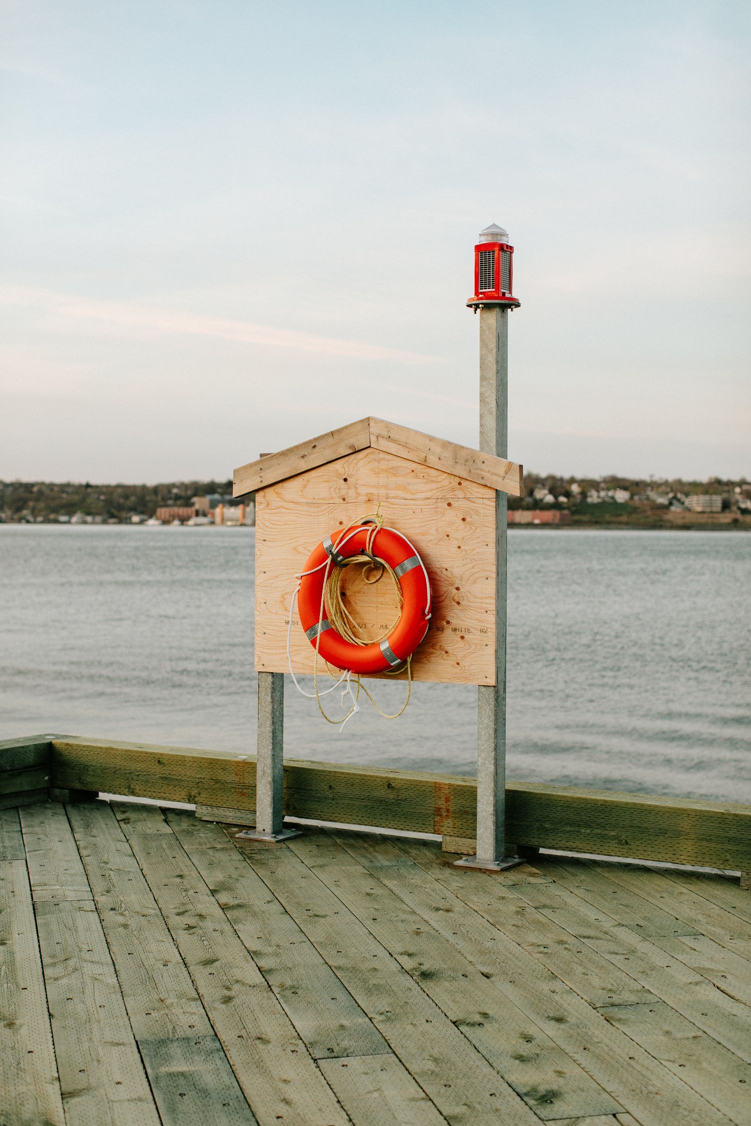 Orange life preserver ring mounted on wooden dock safety station beside red beacon light over calm water.