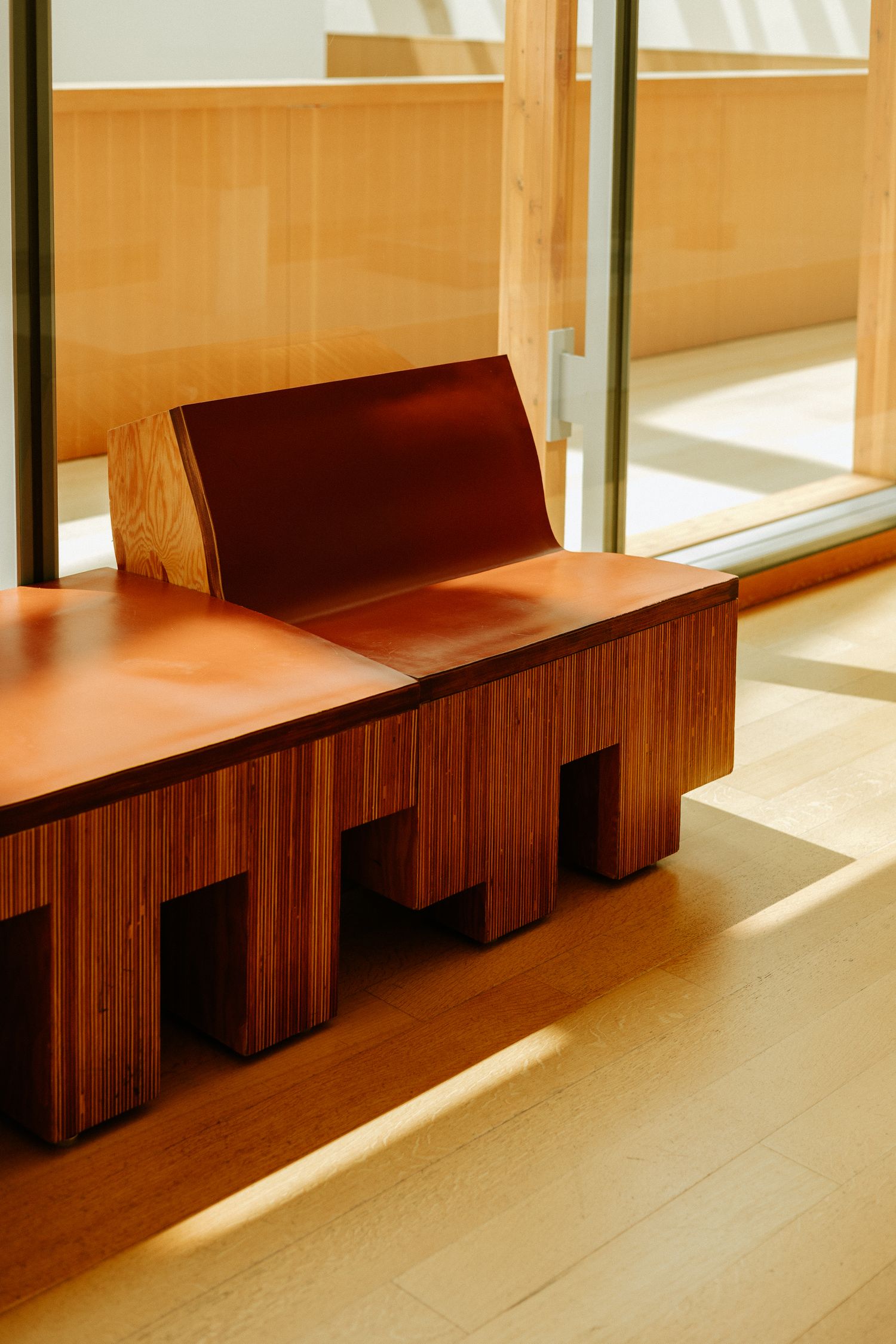 Sculptural wooden bench with curved backrest and geometric cut-out base in warm sunlit interior.