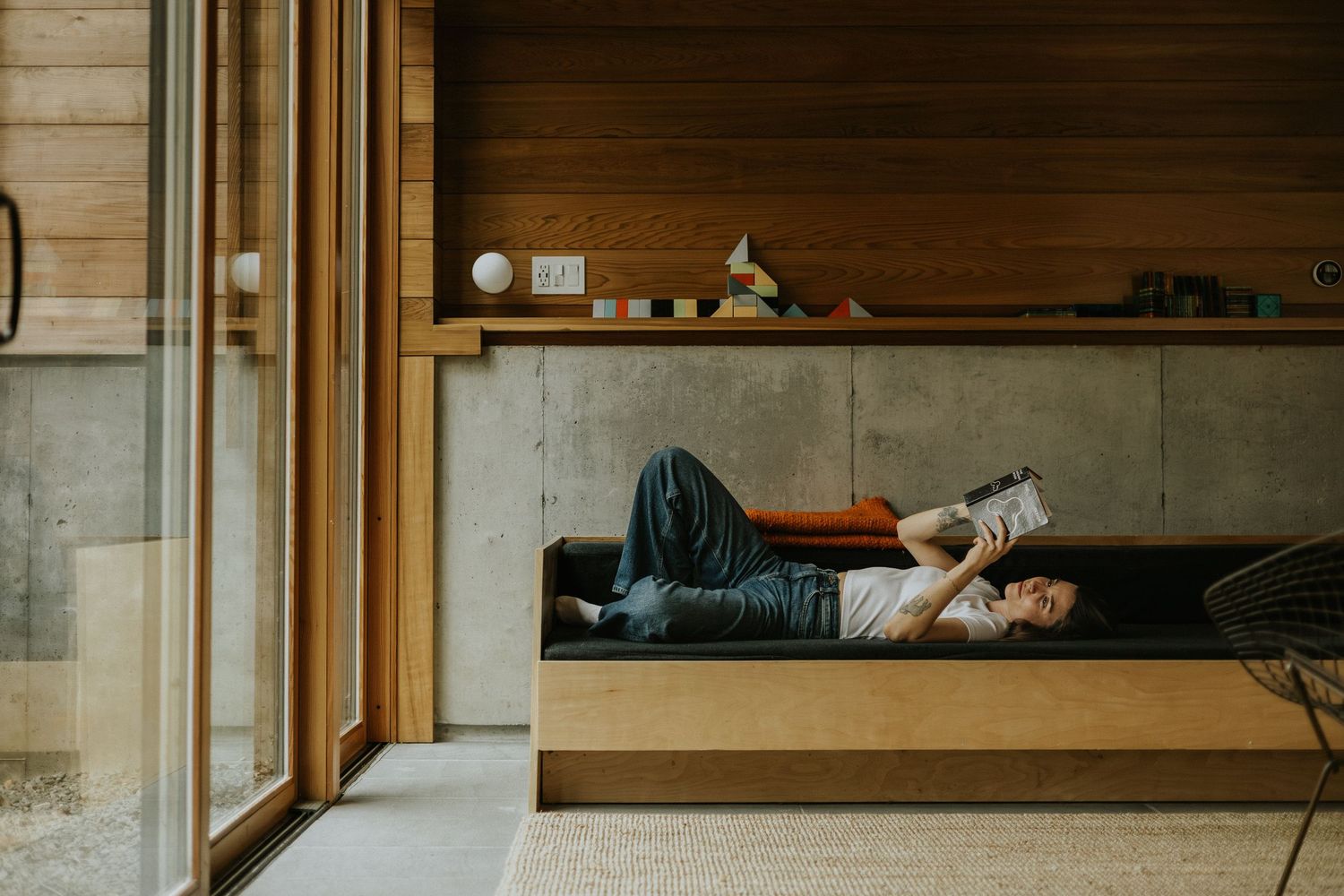 Person relaxing on a modern wooden built-in sofa in a contemporary living room with concrete walls.