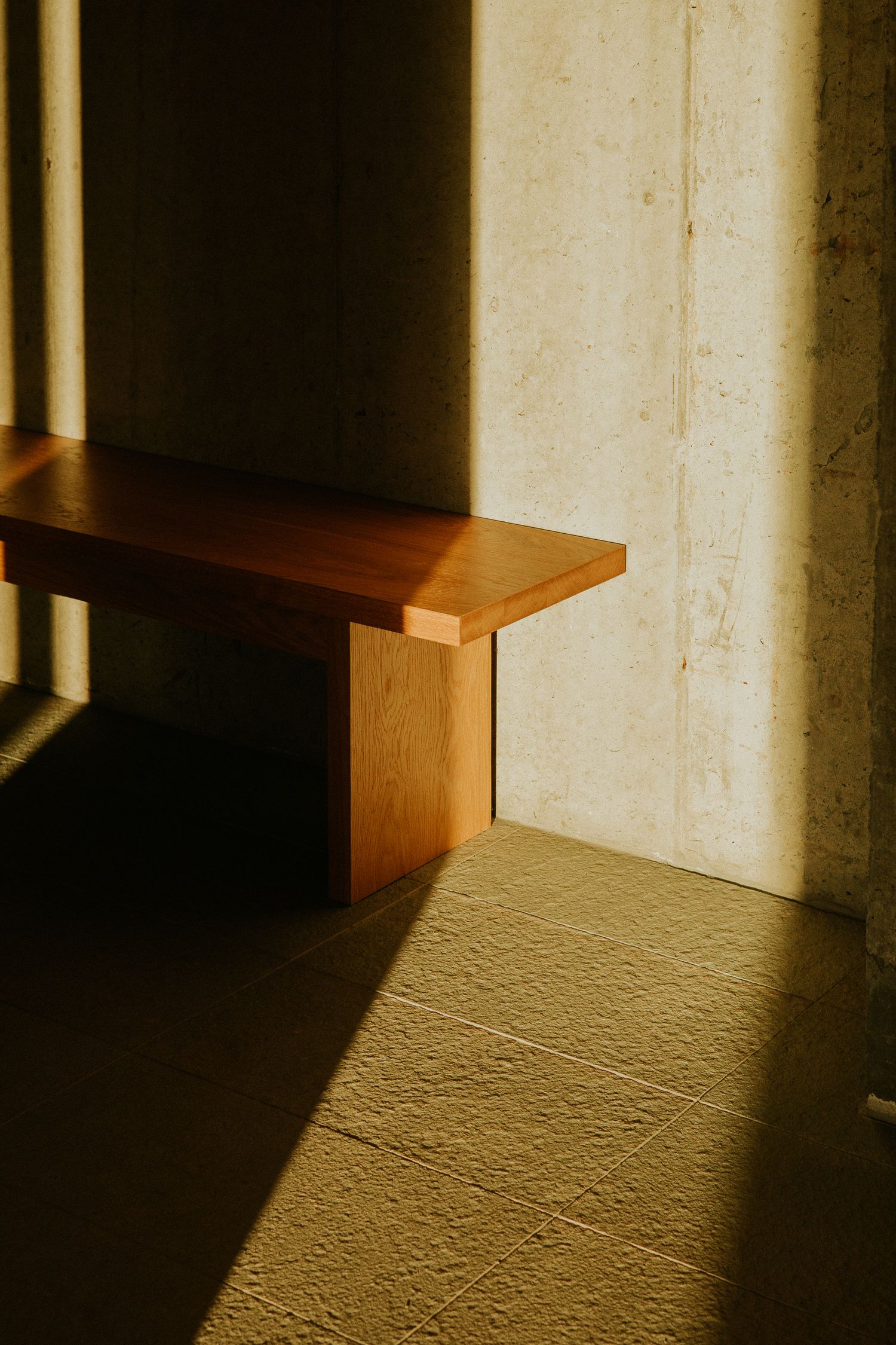 A minimalist wooden bench bathed in warm sunlight against a concrete wall with tiled floor.