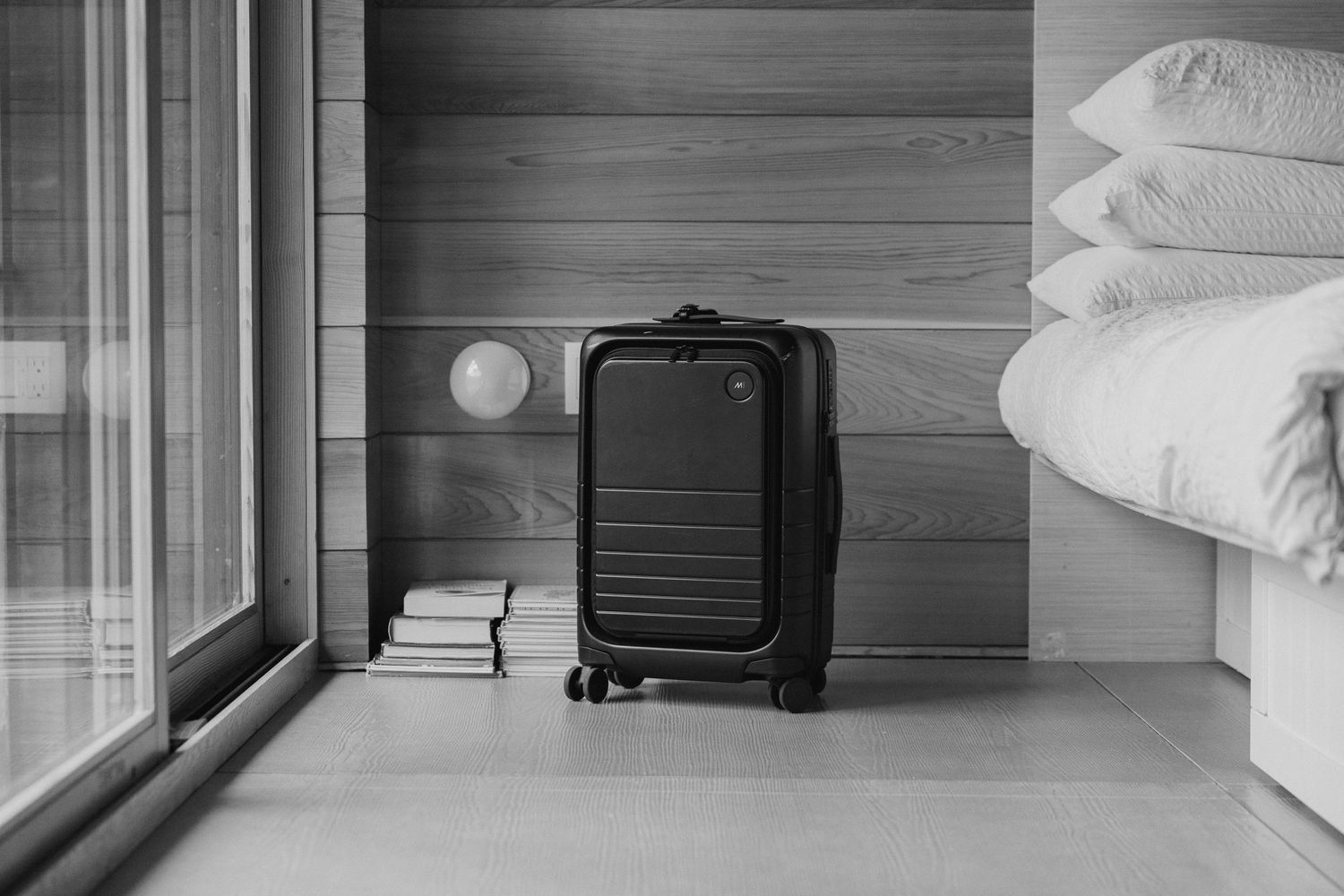 Black hard-shell carry-on luggage standing beside hotel bed with white pillows in monochrome room.