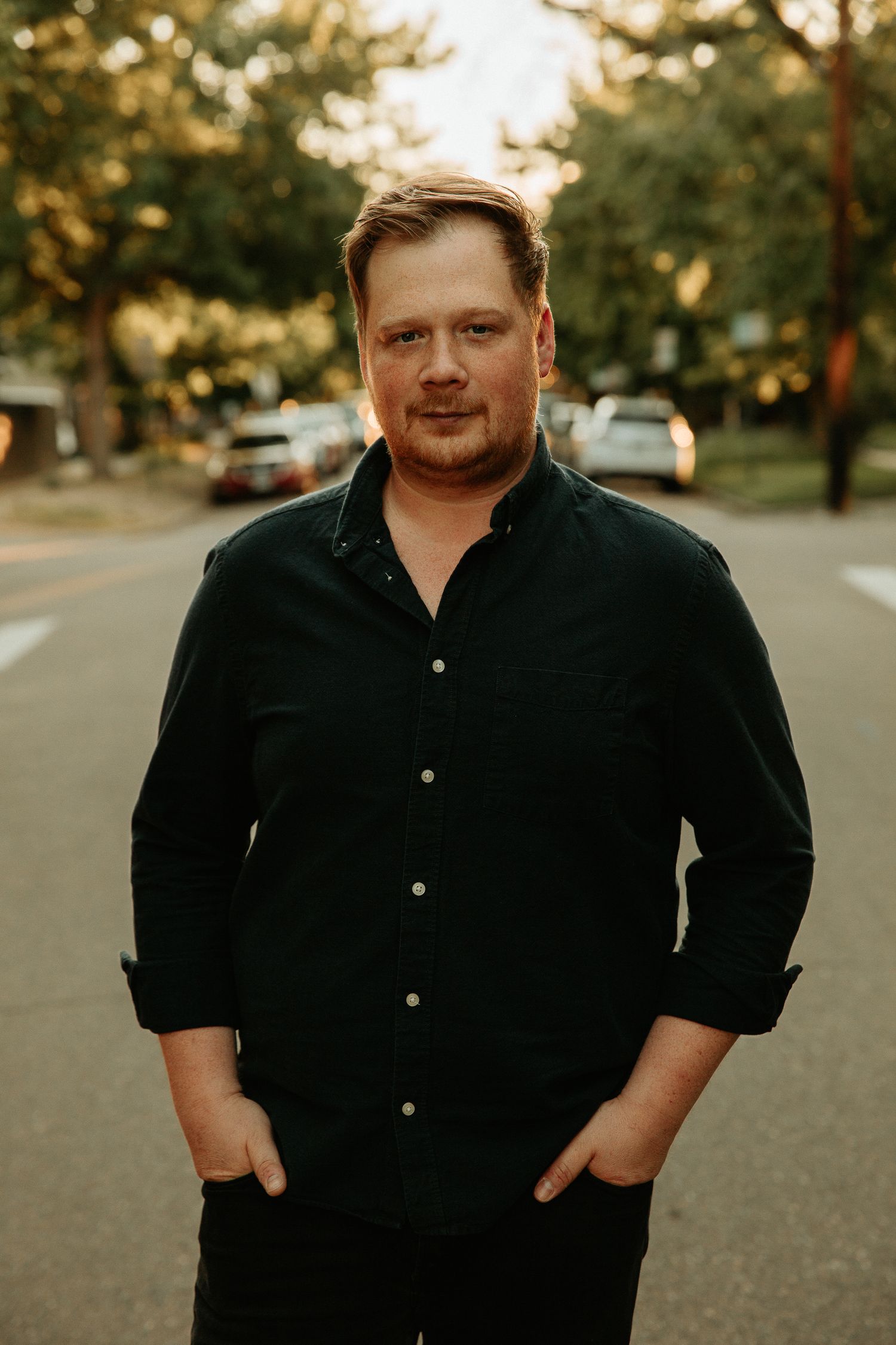 Man in dark button-up shirt standing on a tree-lined residential street at golden hour.
