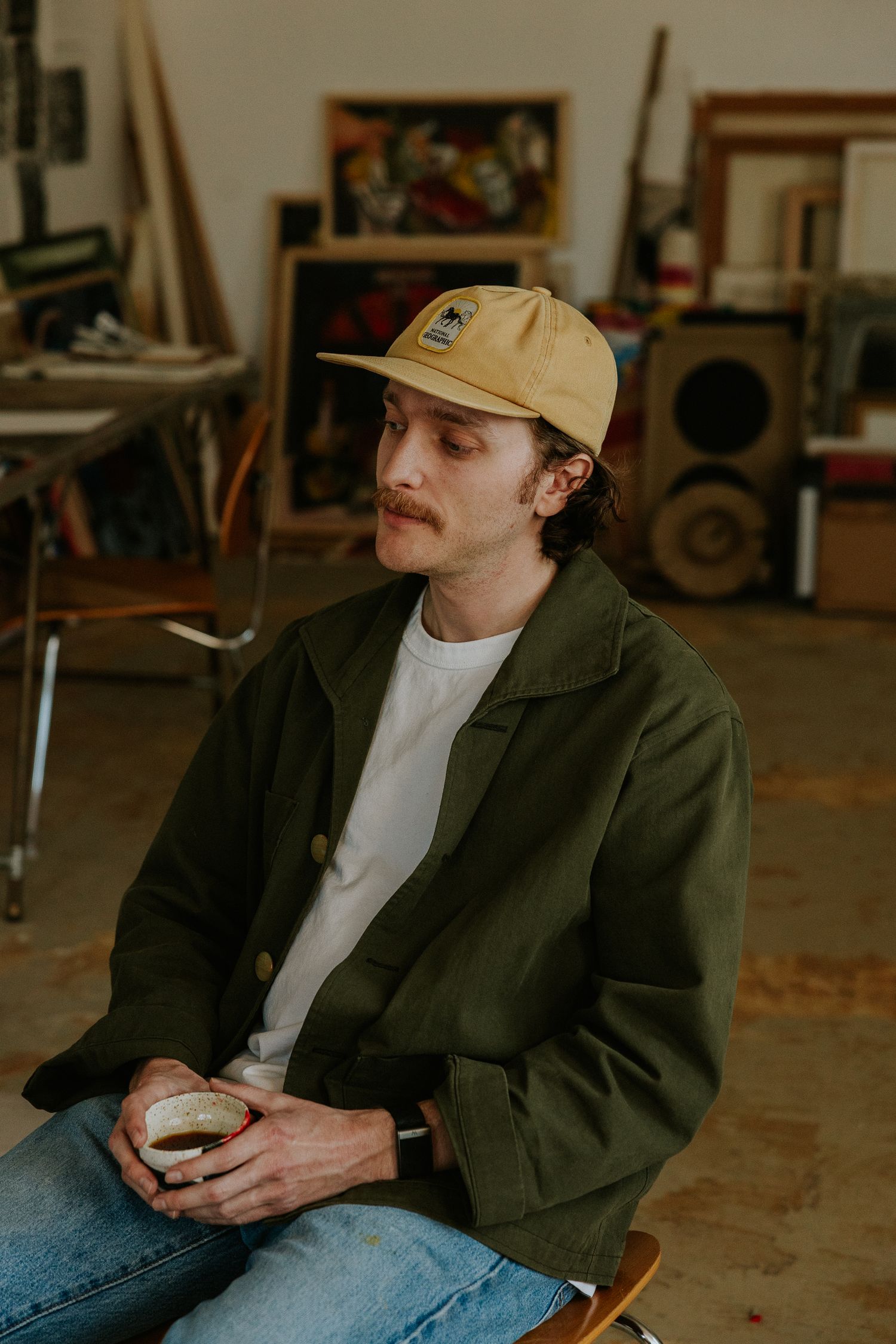 Young man with mustache wearing green jacket and yellow cap holds coffee cup in an art studio.