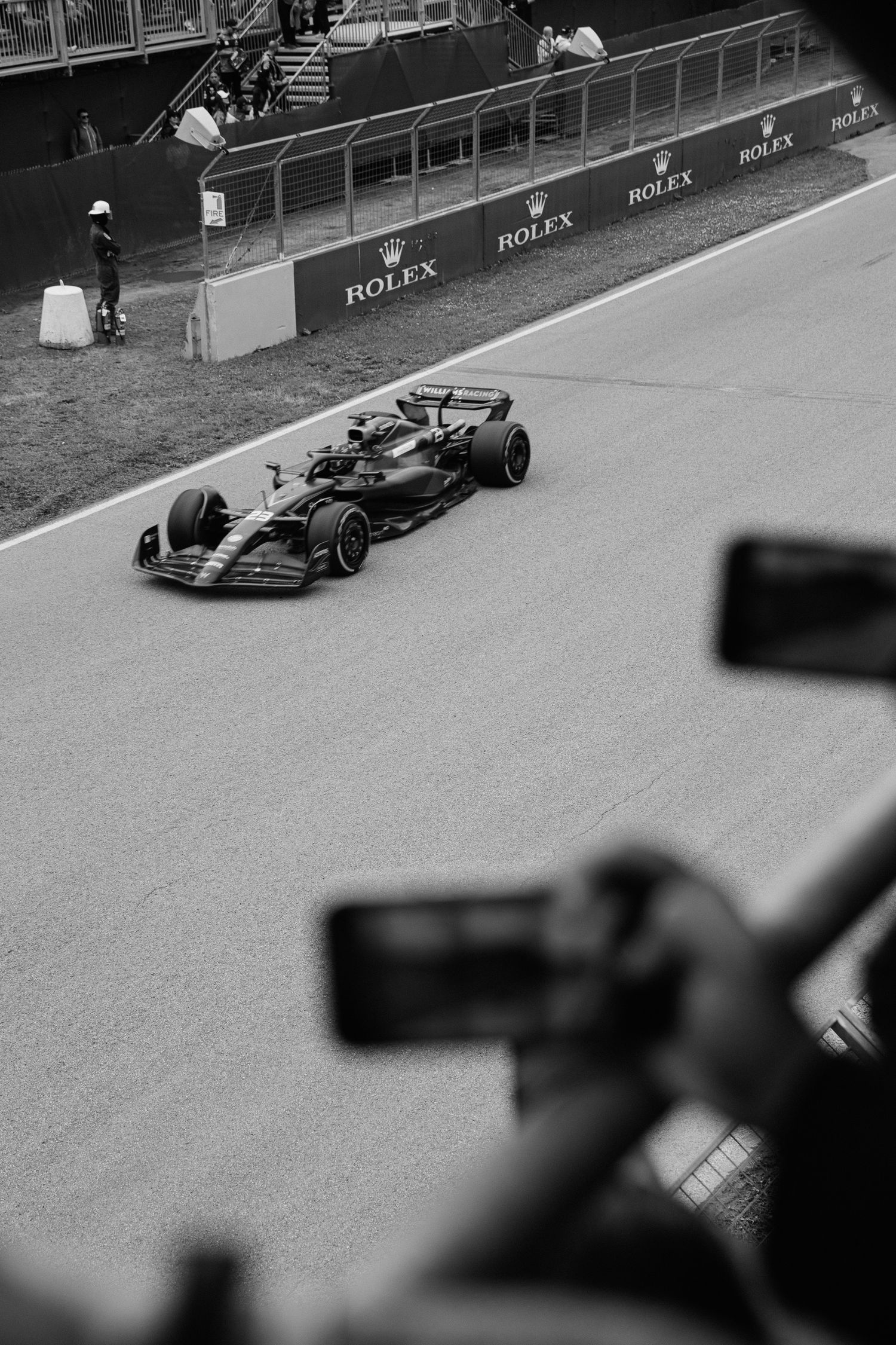 Black and white photo of a Formula 1 race car on track with spectators filming in foreground.