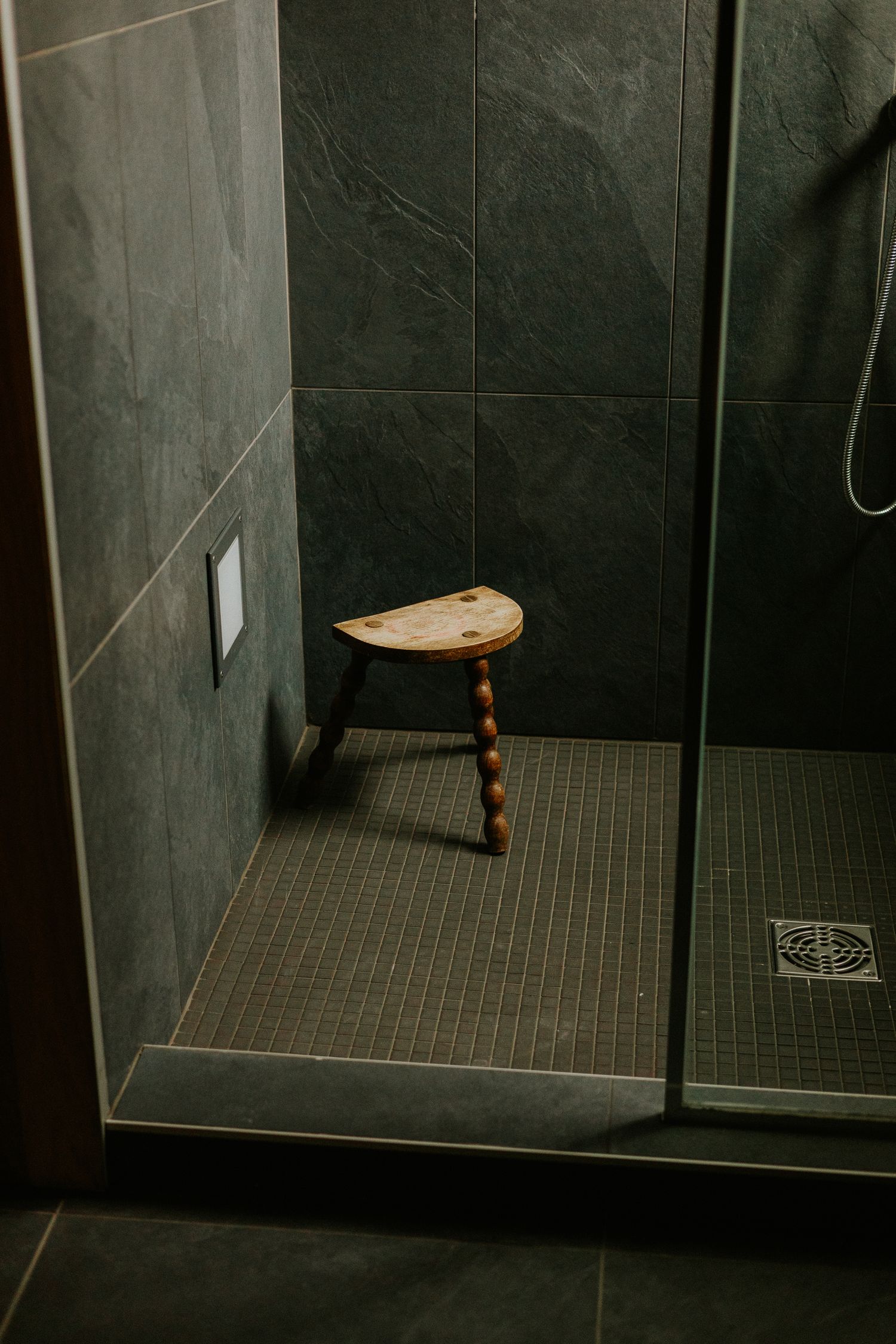 A wooden three-legged stool inside a modern walk-in shower with dark slate tiles and glass door.