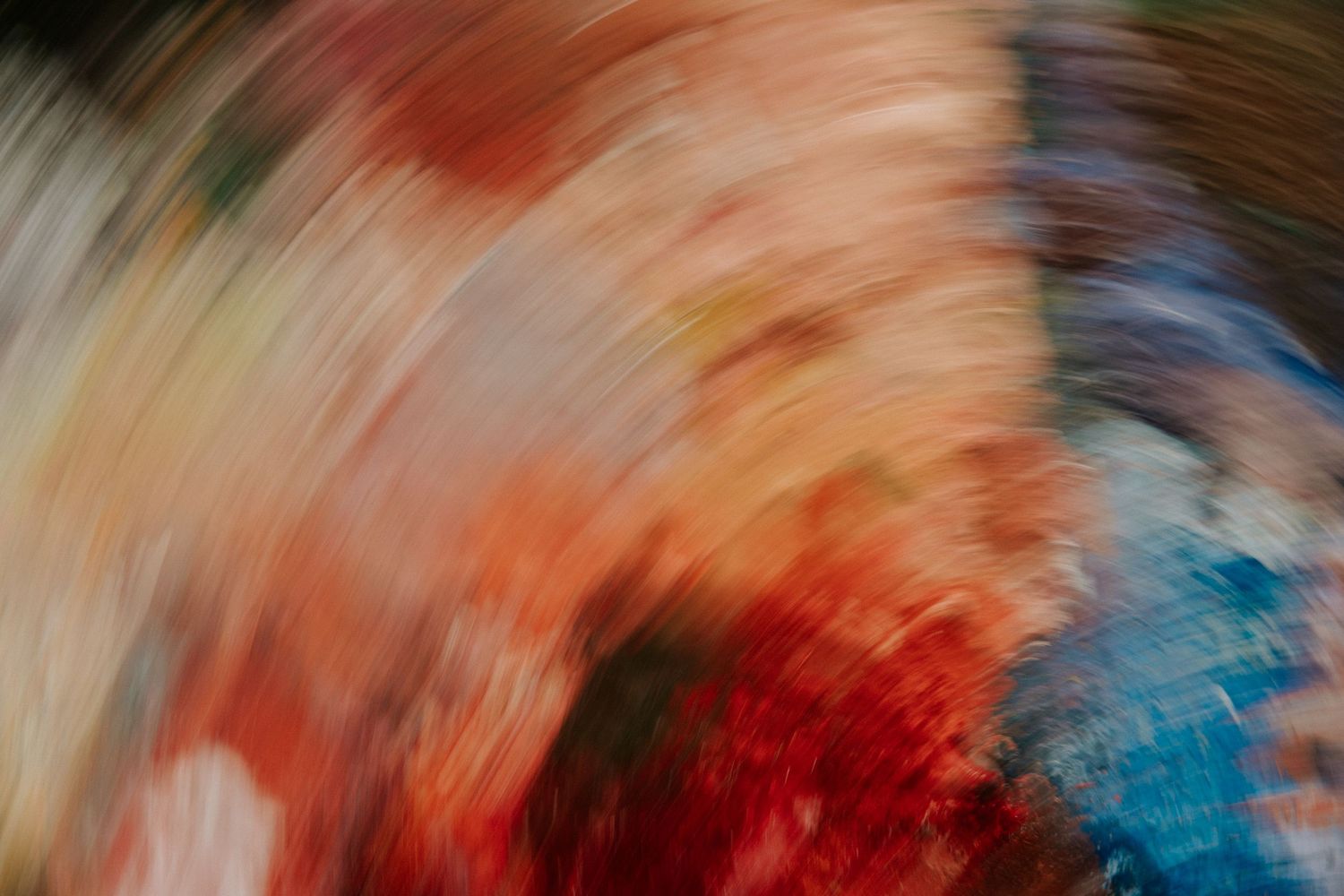 Abstract swirling motion blur art with vibrant red, orange, blue, and cream colors creating a dynamic radial pattern.