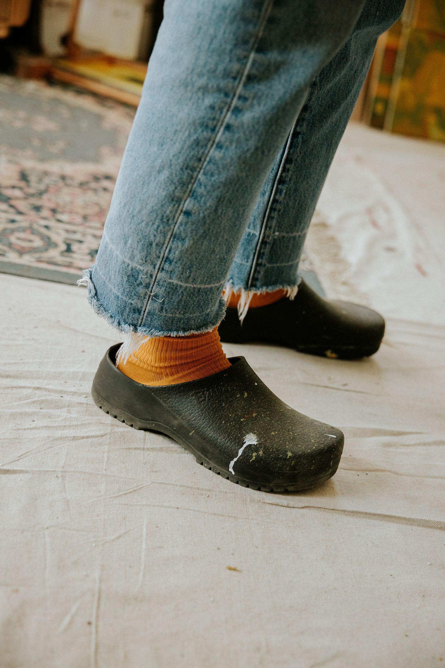Person wearing black clogs with orange socks and frayed hem jeans standing on white drop cloth.