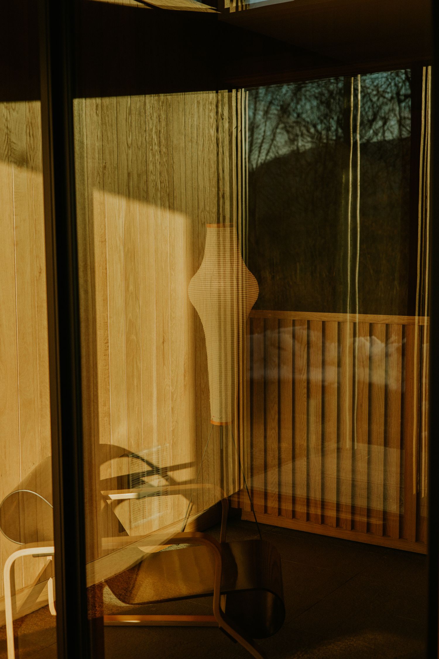 Warm golden sunlight casts a silhouette through sheer curtains onto a cozy balcony with wooden chairs.