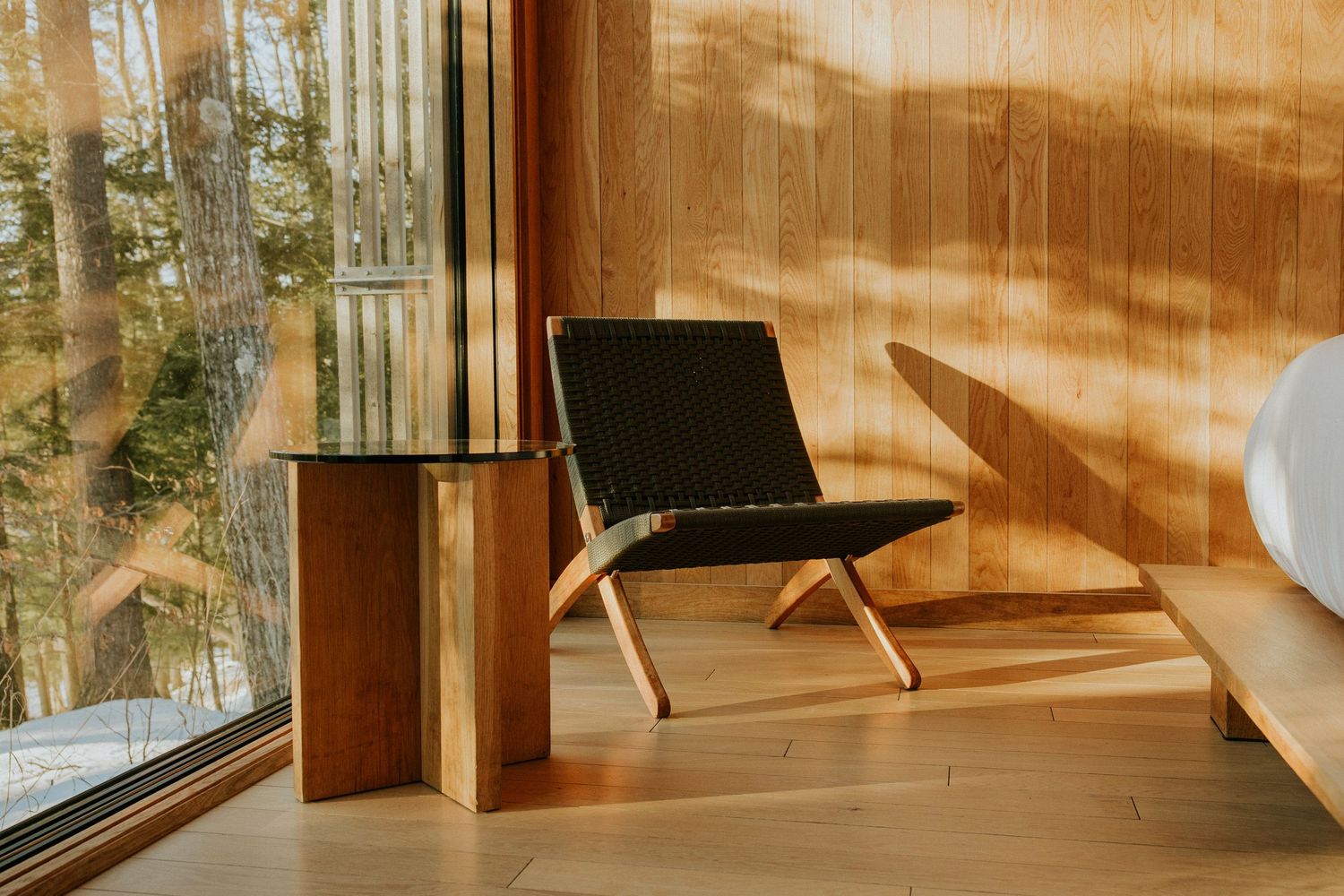 Minimalist interior corner with wood-paneled walls, black woven lounge chair, cylindrical side table, and large forest-view window.