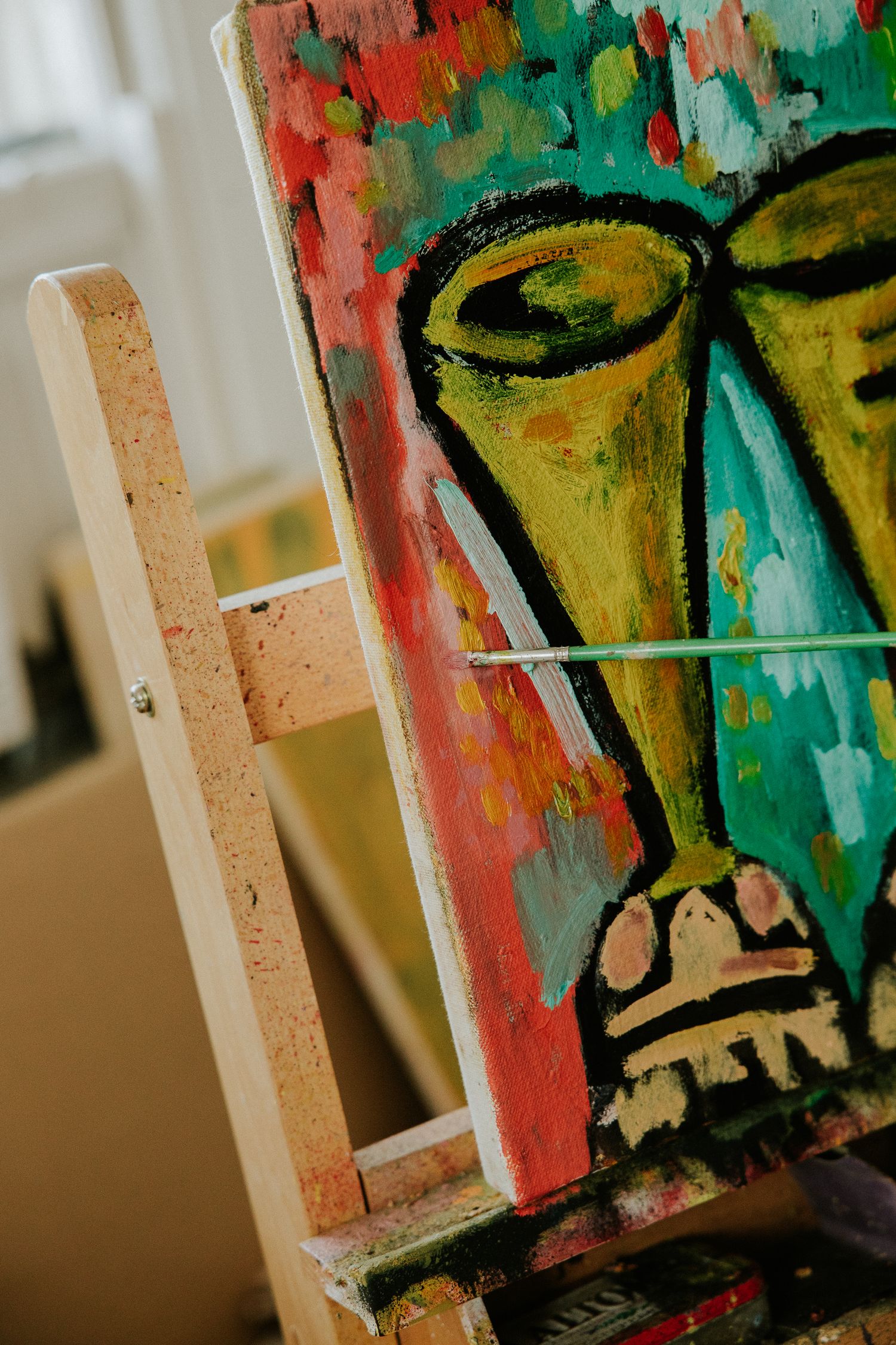 Colorful abstract face painting on canvas easel in art studio, vibrant acrylic artwork with green and red tones.