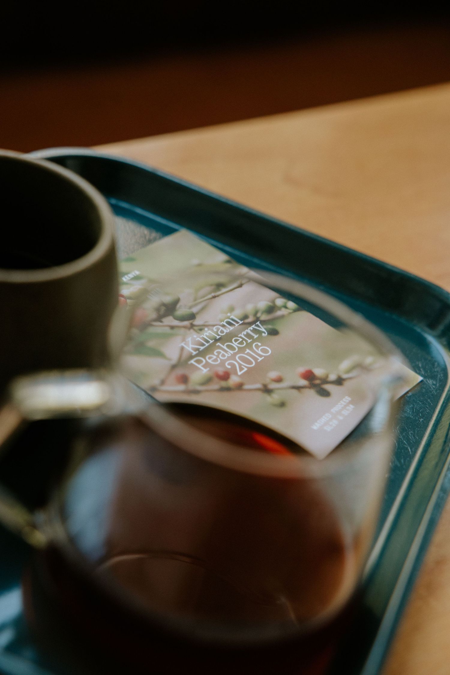 Glass teapot and mug on a teal tray with a Kauai Peaberry 2016 coffee card on a wooden table.