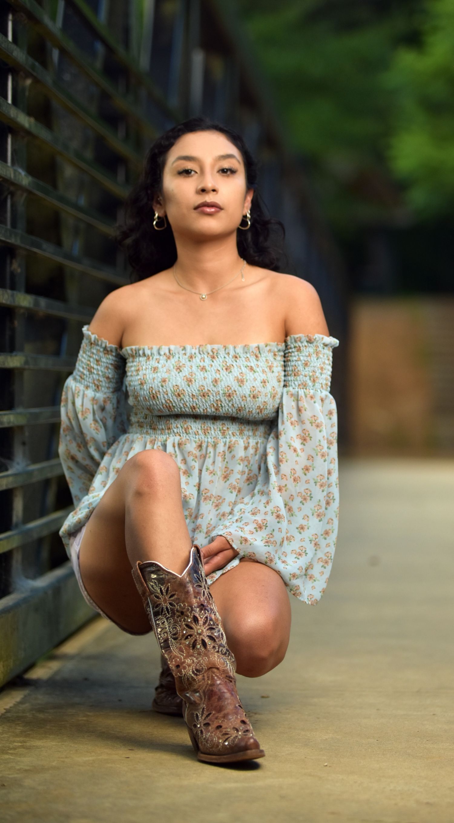 Young woman in floral off-shoulder mini dress and brown cowboy boots kneeling on bridge path.
