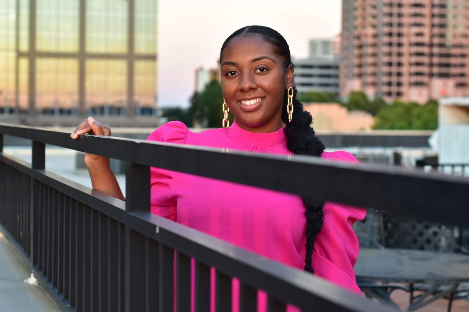 Smiling Black woman in bright pink dress posing on urban rooftop bridge at sunset.