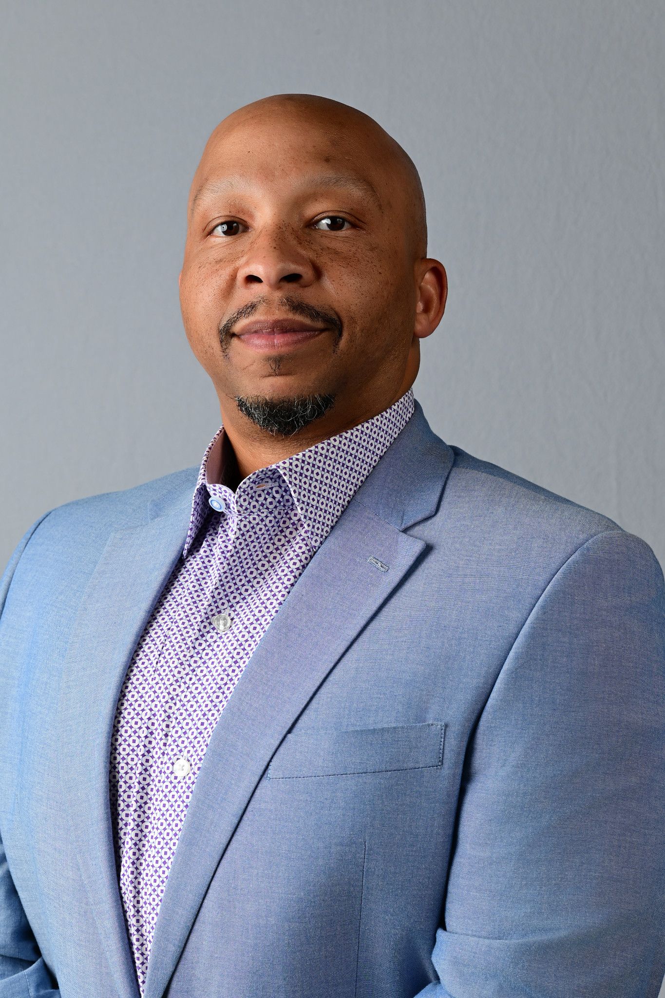 Professional headshot of a bald Black man in a light blue blazer and patterned shirt, smiling confidently.