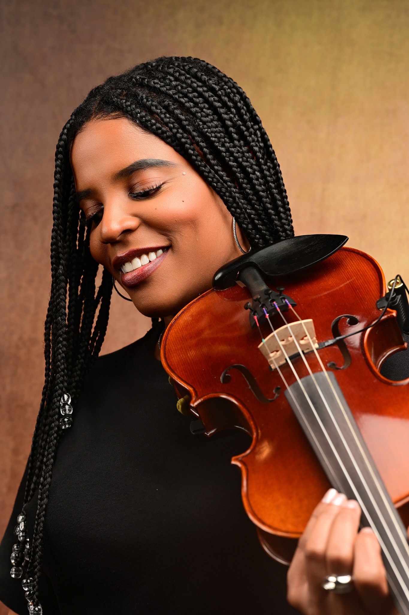 Smiling violinist with long braids and closed eyes rests her cheek on a polished wooden violin in a warm studio portrait.