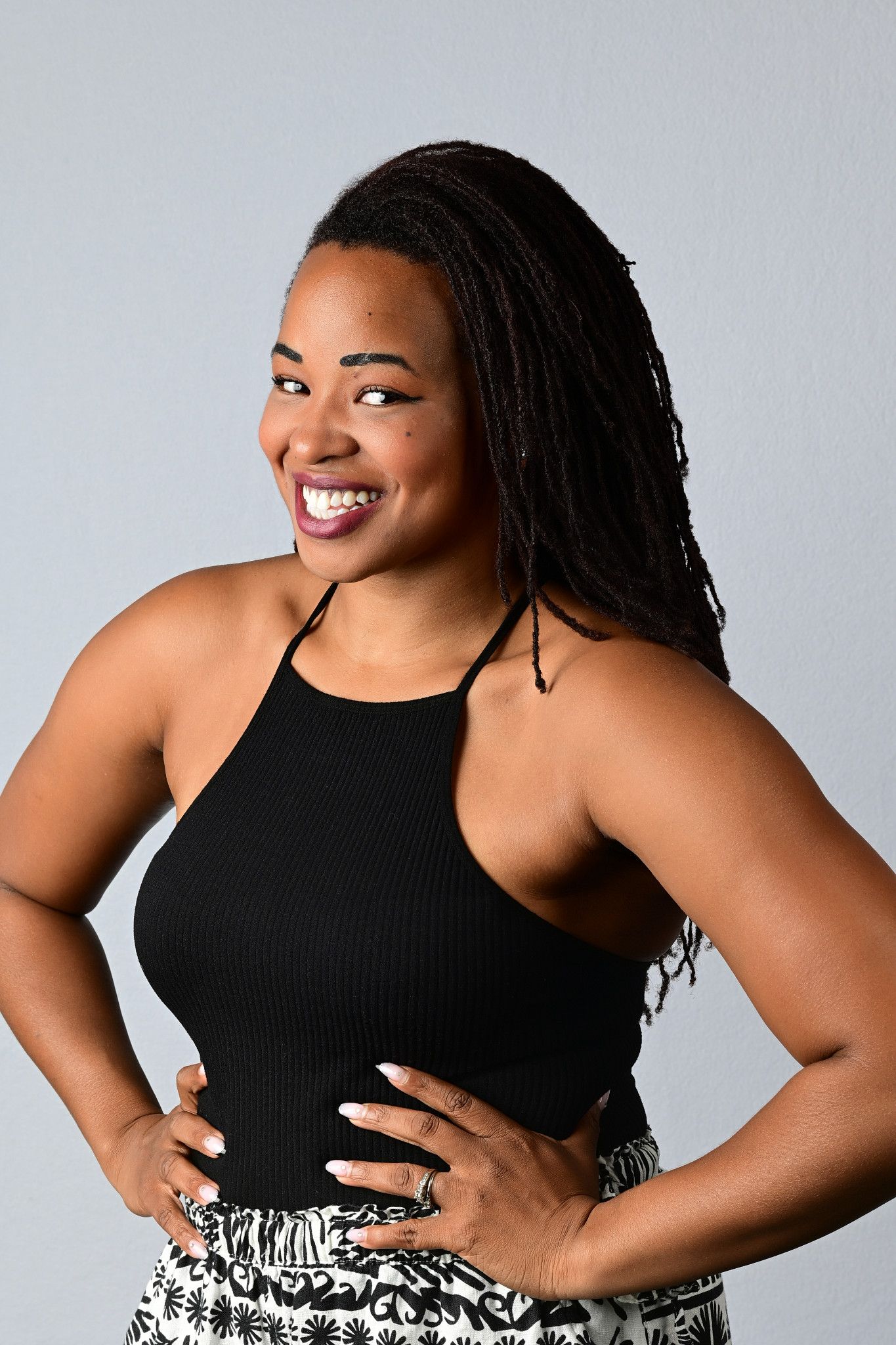 A smiling woman with long braided hair poses with hands on hips in a black halter top against a light gray studio background.